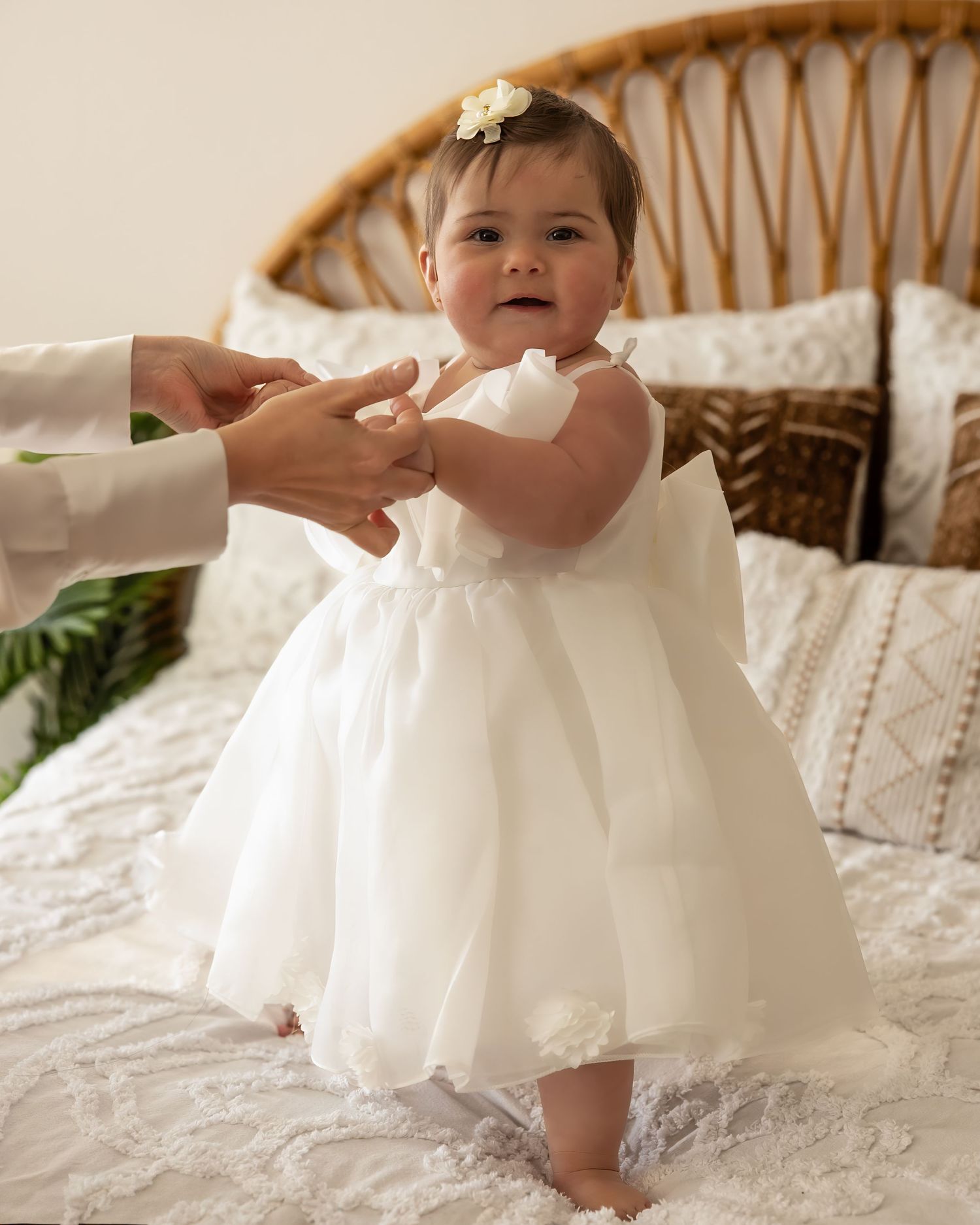 Adorable baby girl in white tulle dress standing on a bed holding an adult's hands for balance.