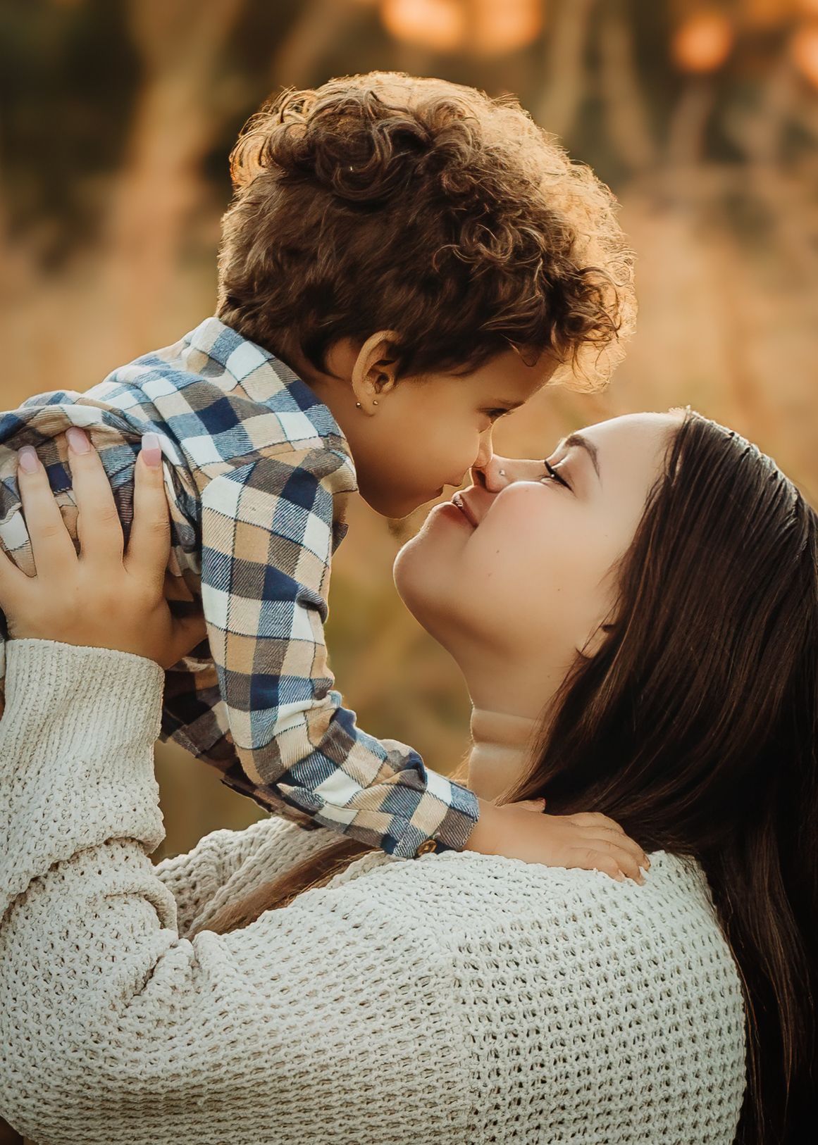 Mother in cream sweater lifting curly-haired toddler for an Eskimo kiss in warm autumn light.