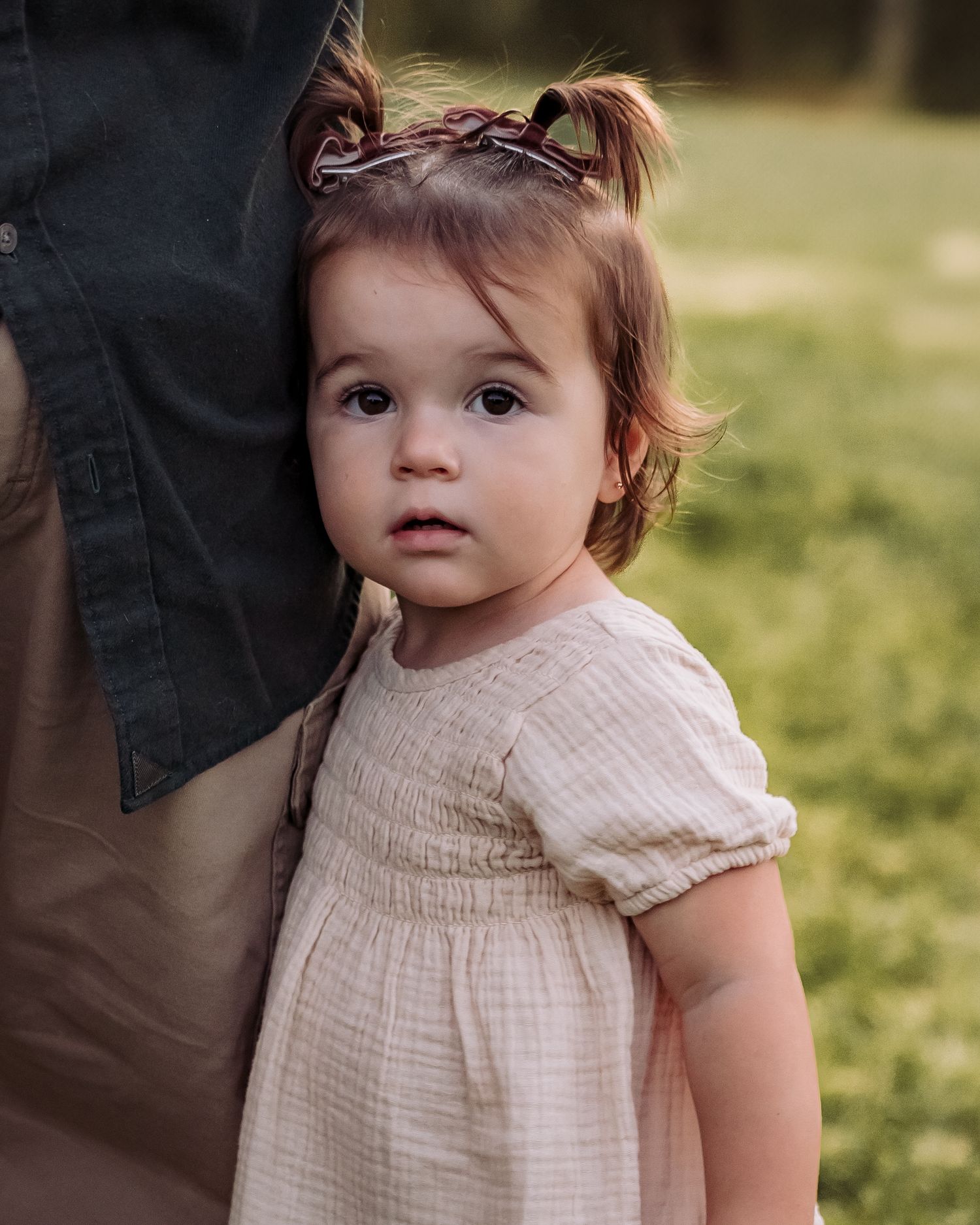 A toddler girl in a cream dress with pigtails leans against an adult, looking curiously at the camera outdoors.