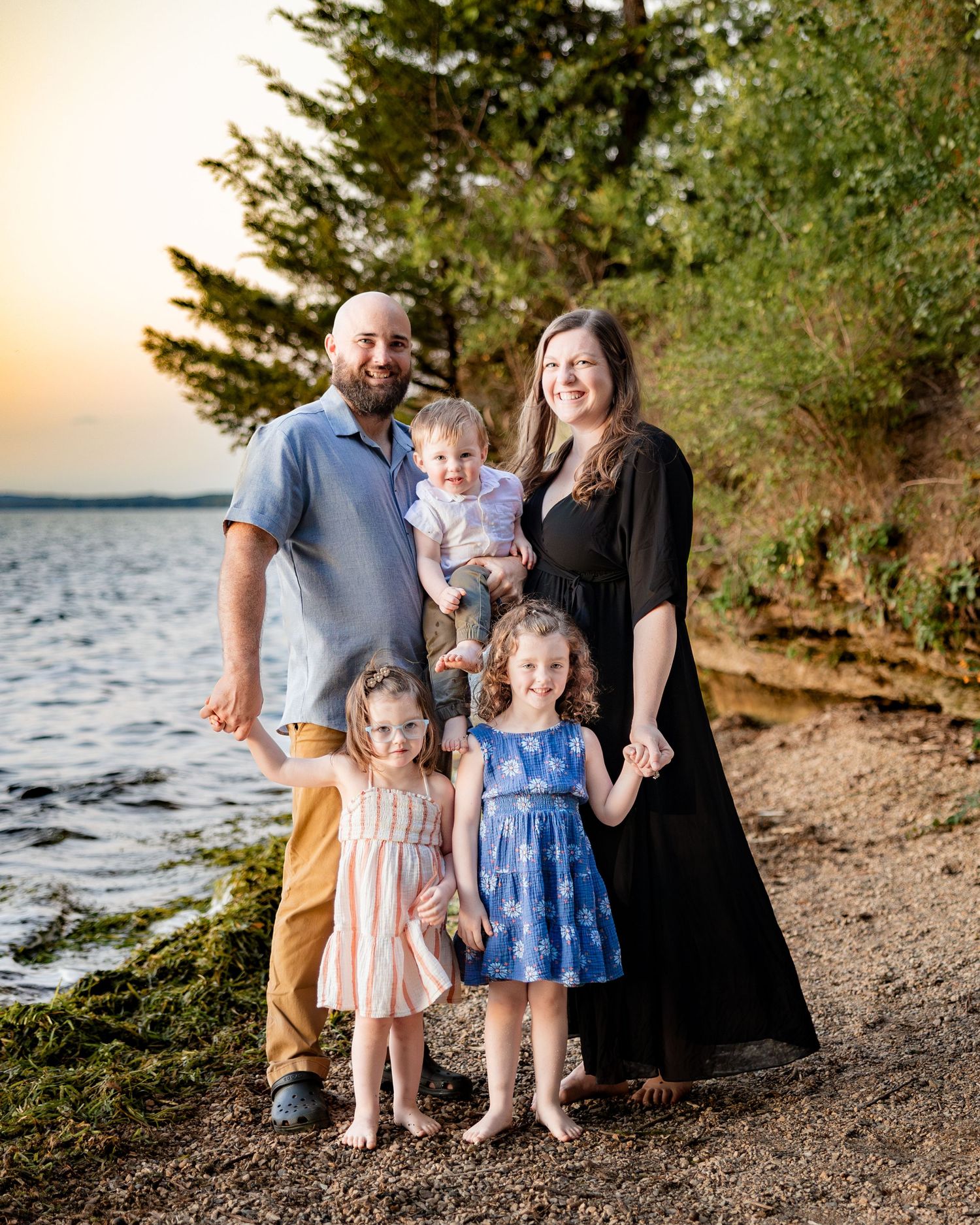 Family of five posing on a rocky beach at sunset, with tall trees and calm water in the background.