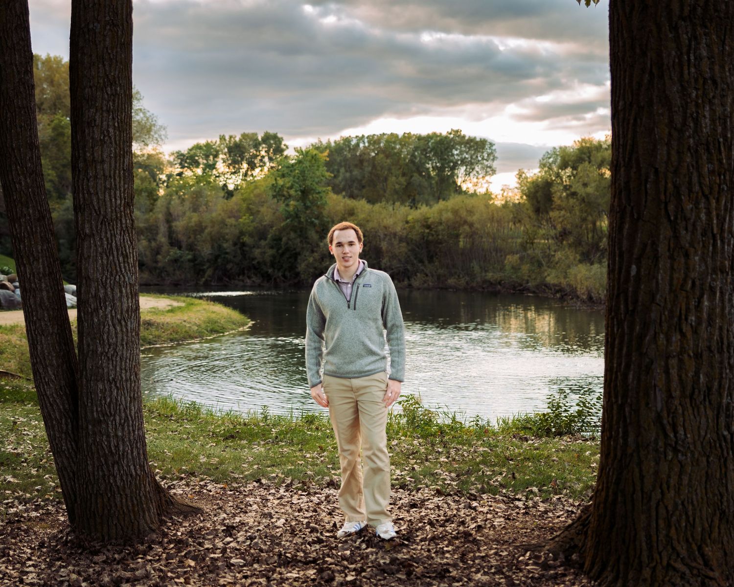 Man in gray hoodie and khaki pants stands between two trees near a serene river with lush greenery under cloudy skies.