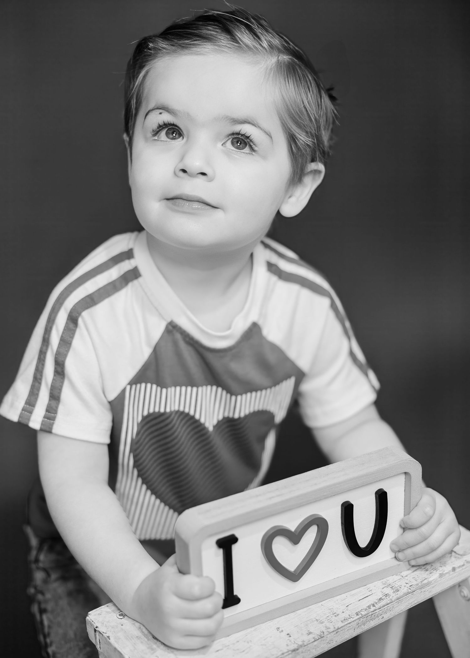 Black and white portrait of toddler boy holding I Love You sign while seated on chair.