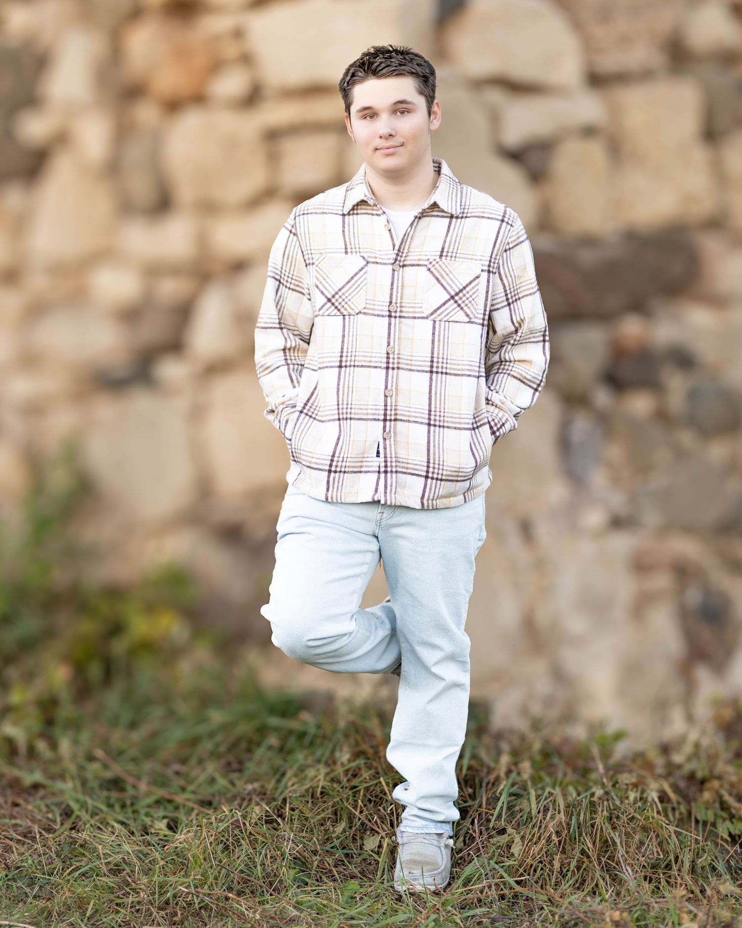 Teen boy in plaid flannel shirt poses against rustic stone wall for senior photos.