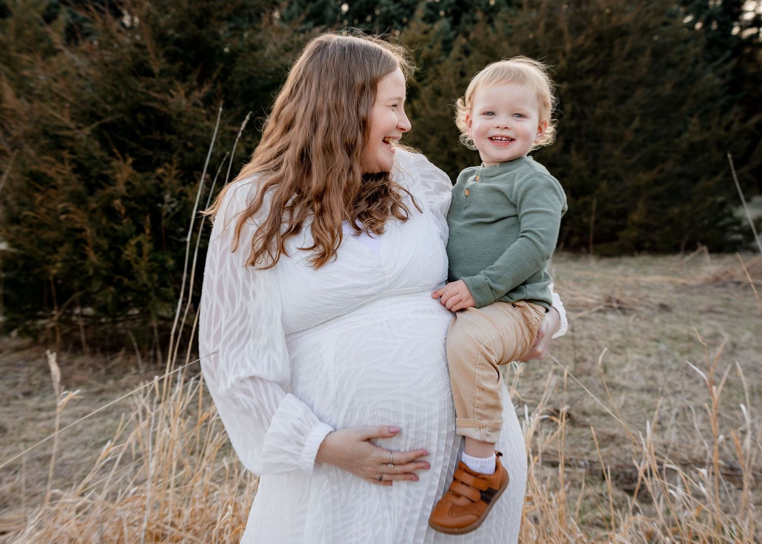 A pregnant woman in a white dress holds a smiling toddler boy in a field of tall grass.