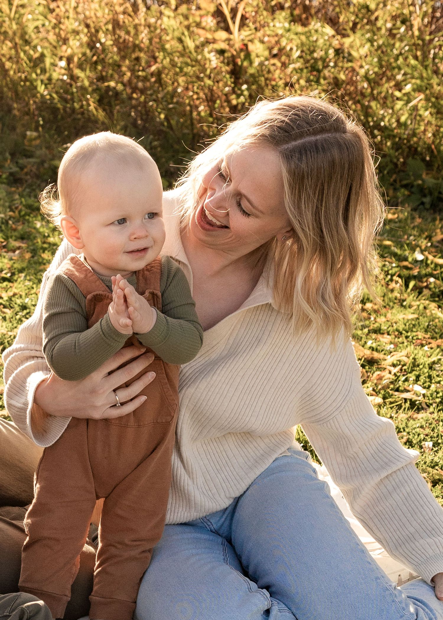 Smiling blonde mother sitting on grass holding her happy clapping baby in warm fall sunlight.