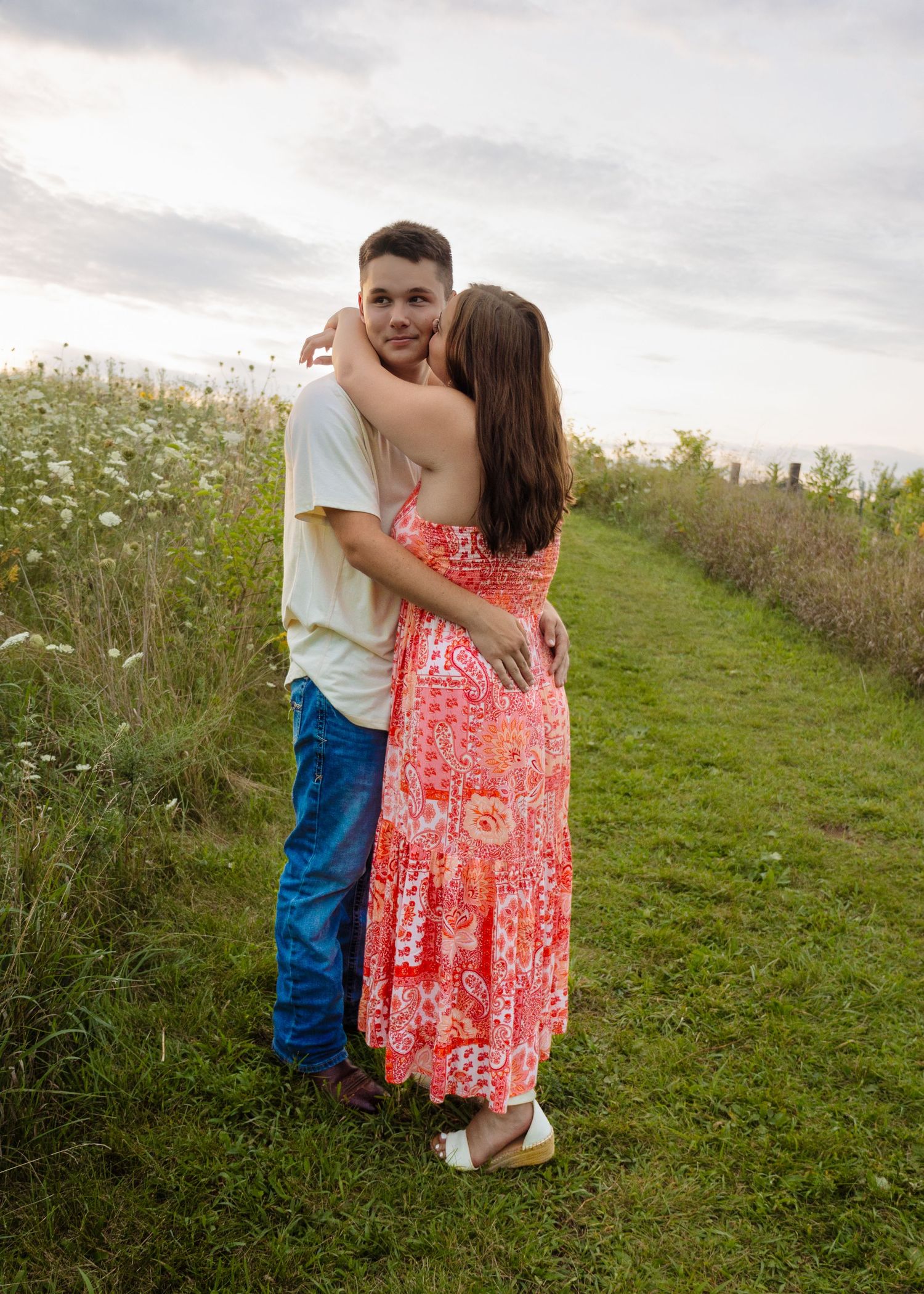 Young couple embracing on a grassy path lined with wildflowers, woman in pink floral maxi dress at sunset.