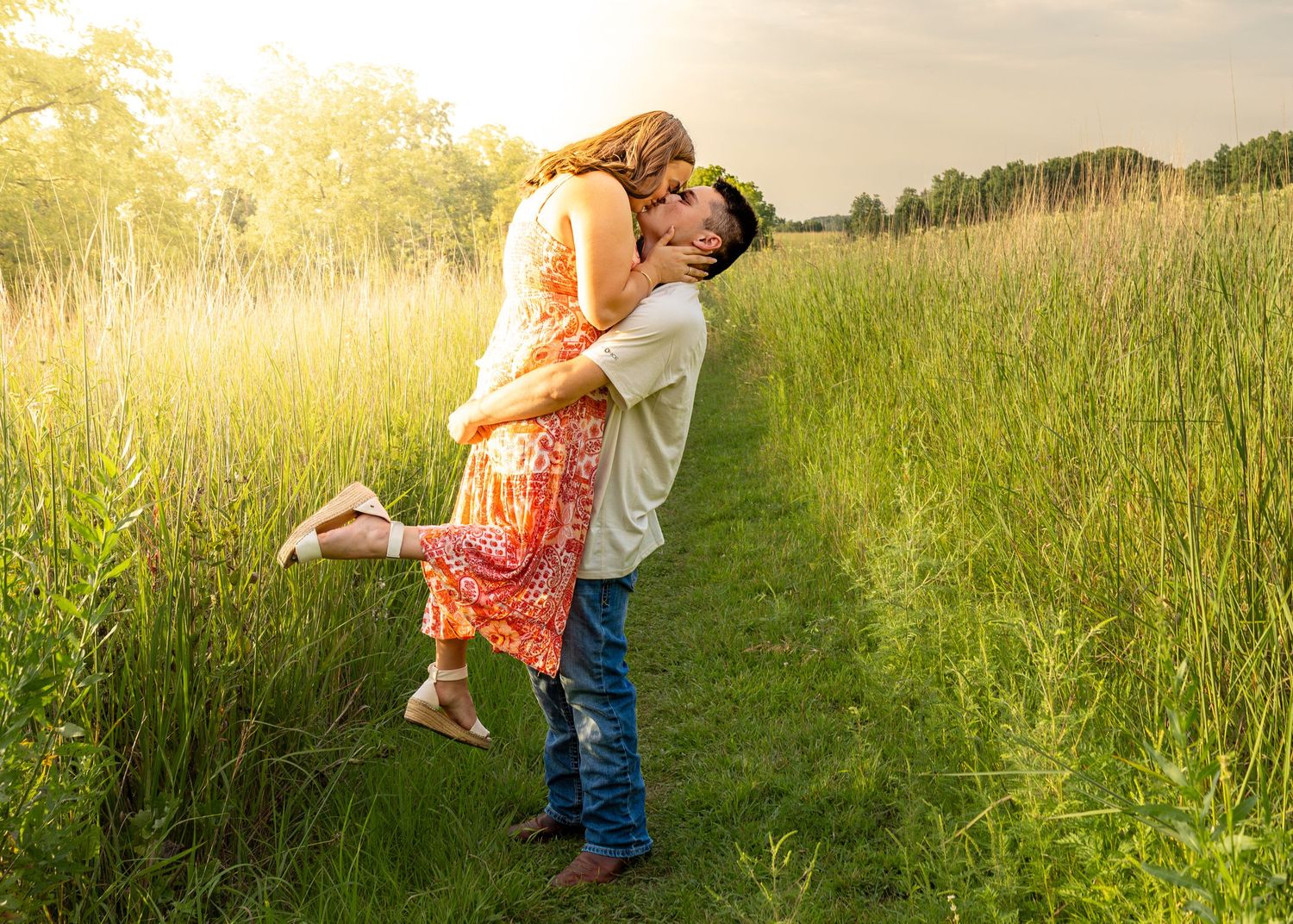 Couple sharing a romantic kiss on a sunlit meadow path, woman lifted off ground in red floral dress.