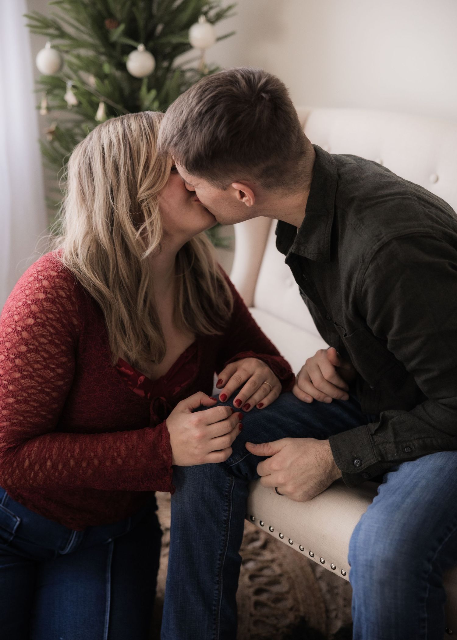 Couple sharing a kiss while seated together near a decorated Christmas tree indoors.