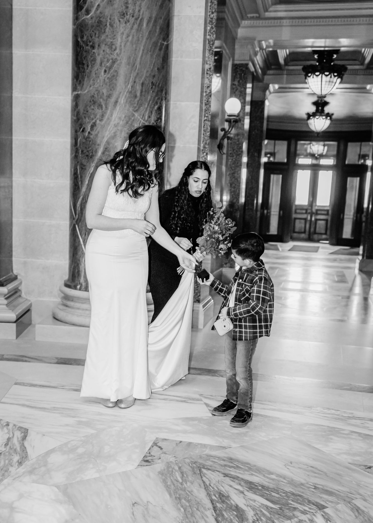 Bride in white gown and guest receiving flowers from a young child in a grand marble hallway, black and white.