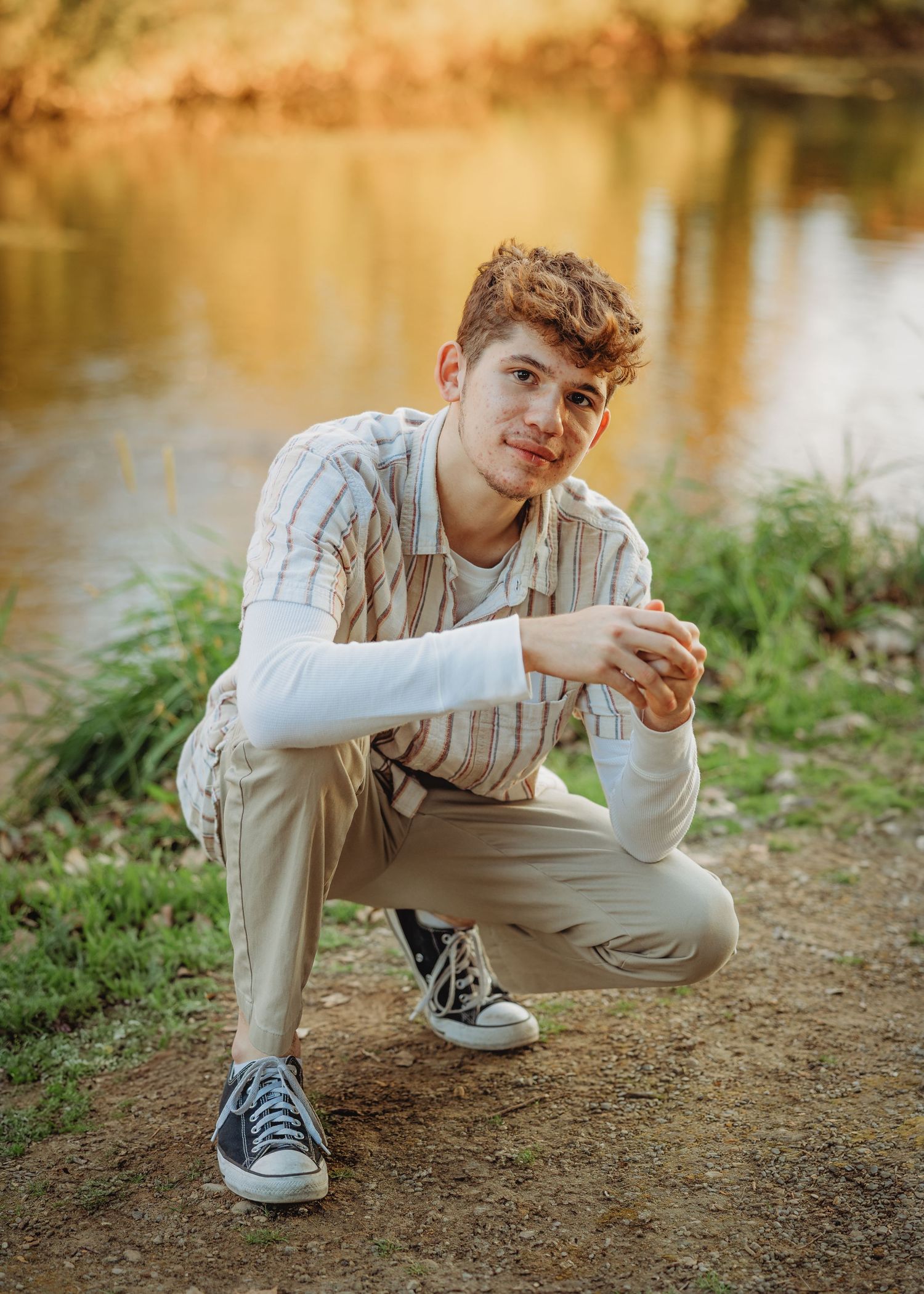 Young man crouching near a serene lake during golden hour in a striped shirt and khaki pants.