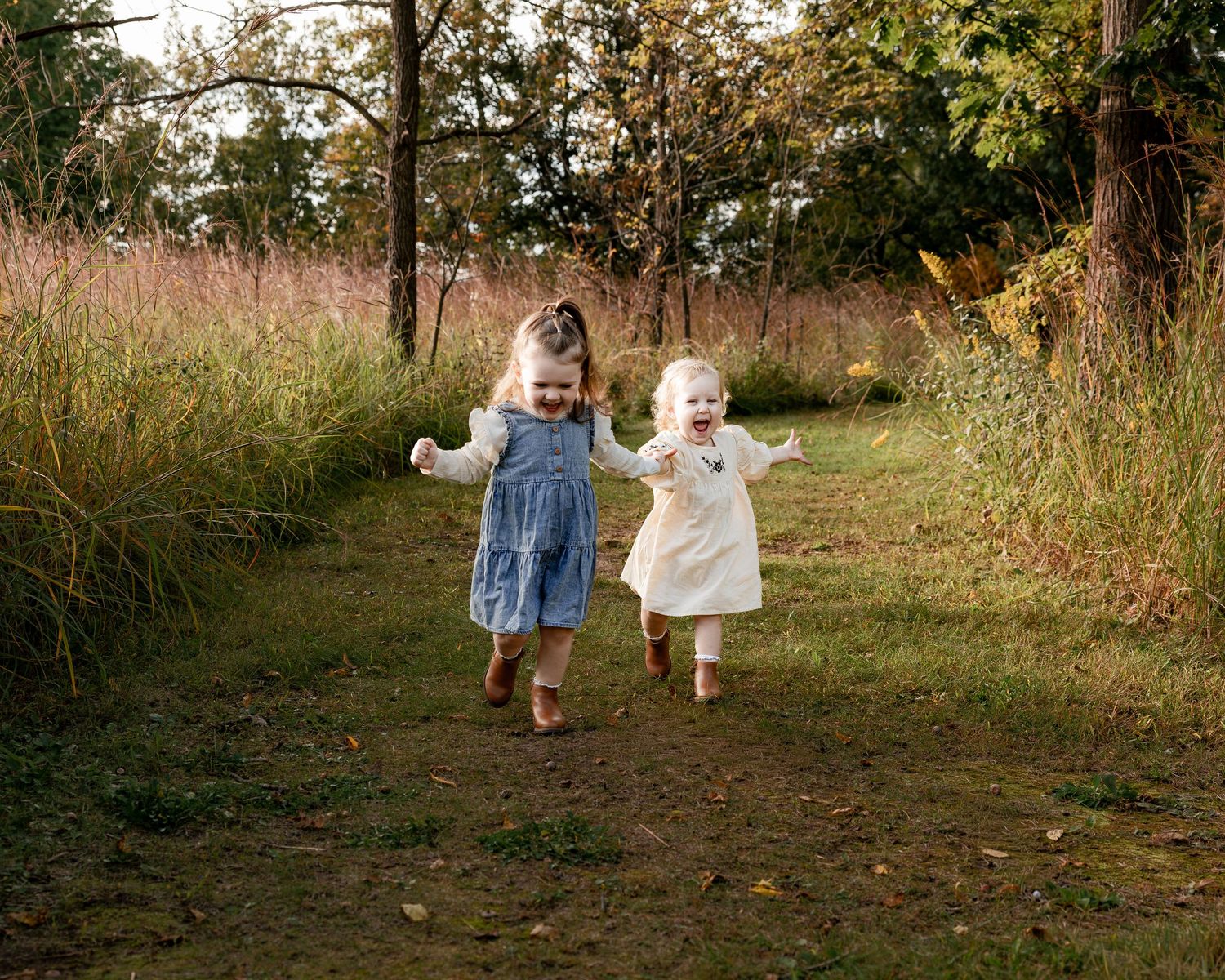 Two young girls in dresses joyfully running on a dirt path through a lush green meadow.