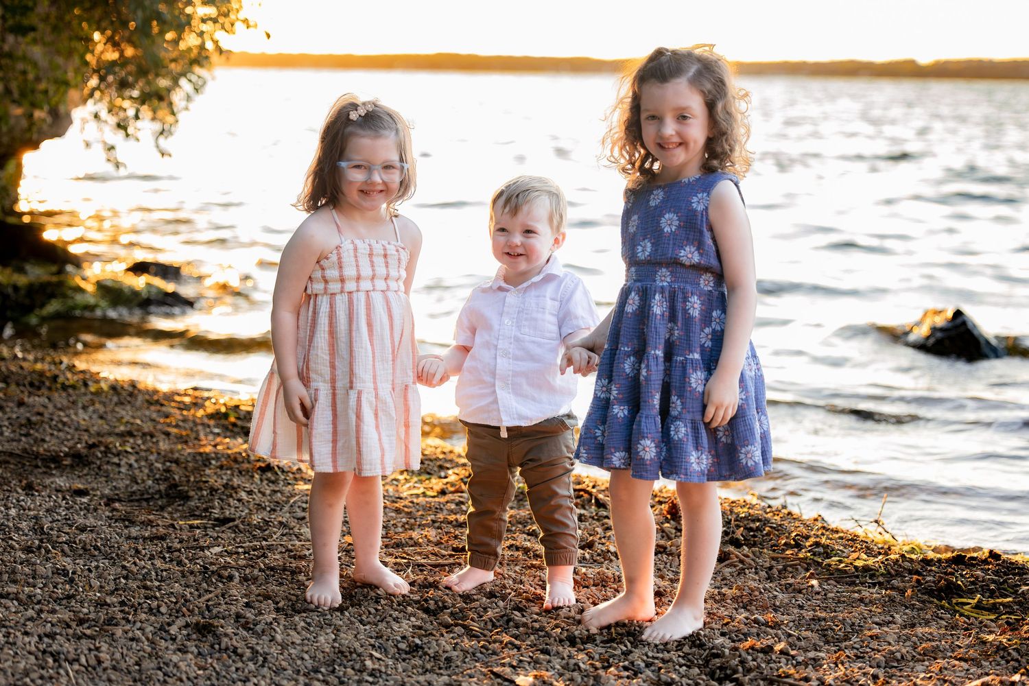 Three young children smiling together on a rocky lakeside beach during golden hour sunset.