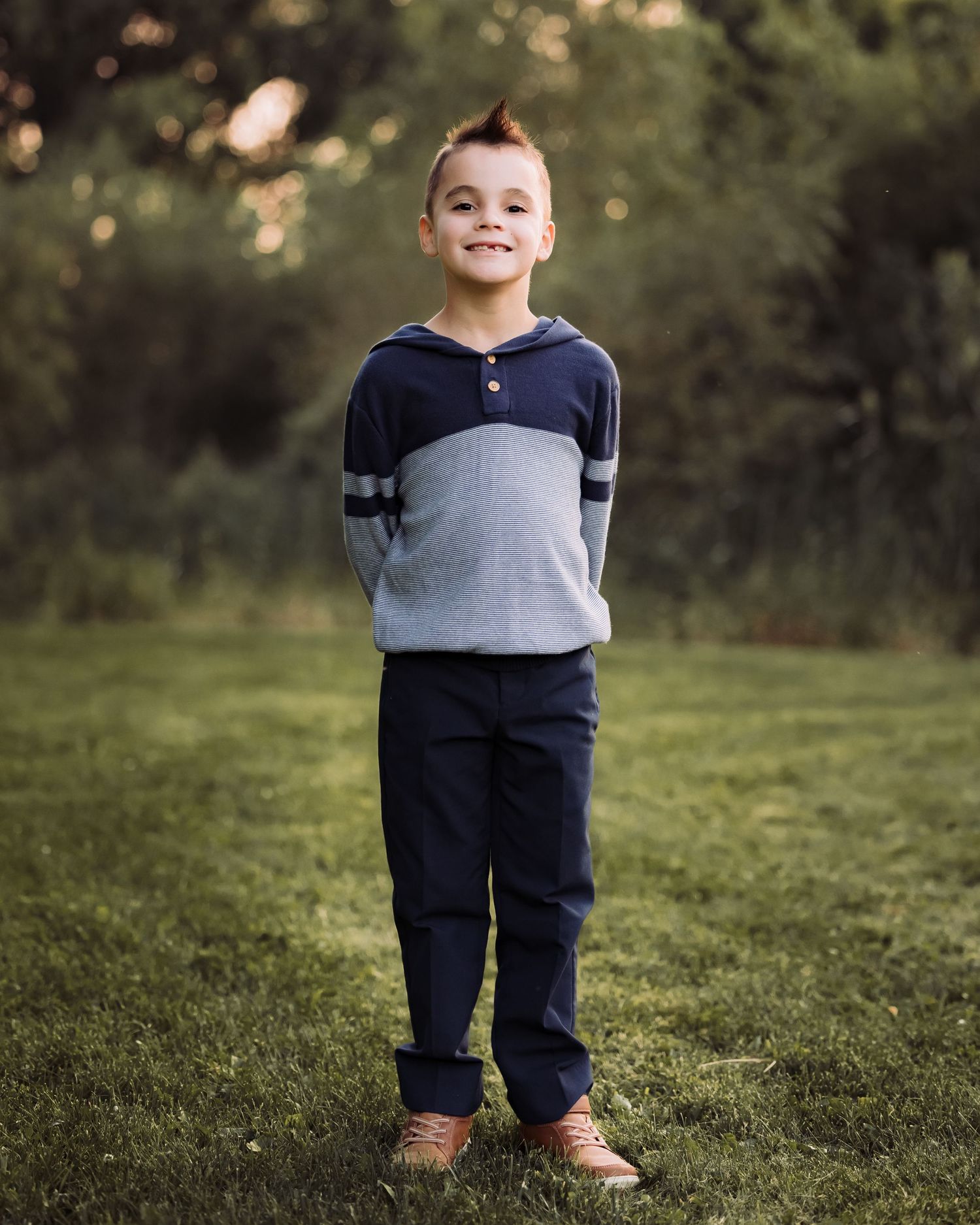 Young boy smiling outdoors in navy striped hoodie and navy pants, standing on green grass with trees in background.