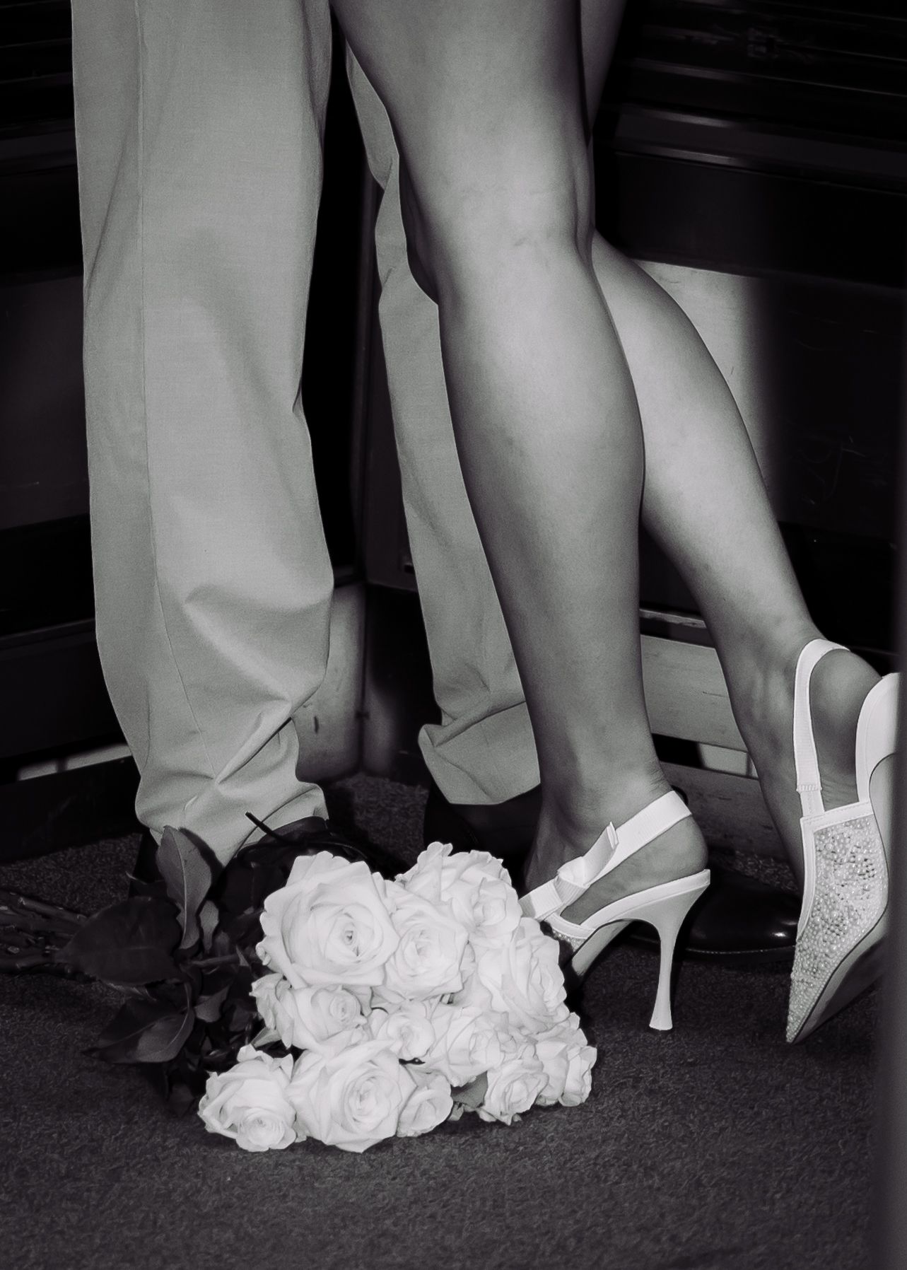 Black and white wedding photo of bride in heels and groom standing beside a white rose bouquet on the floor.