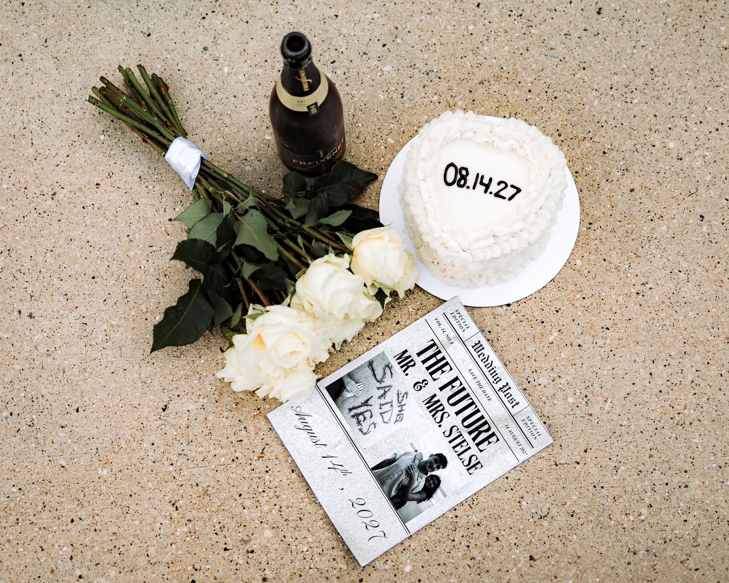 Wedding flat lay with white roses, cake dated 08.14.27, champagne bottle, and newspaper on beige surface.