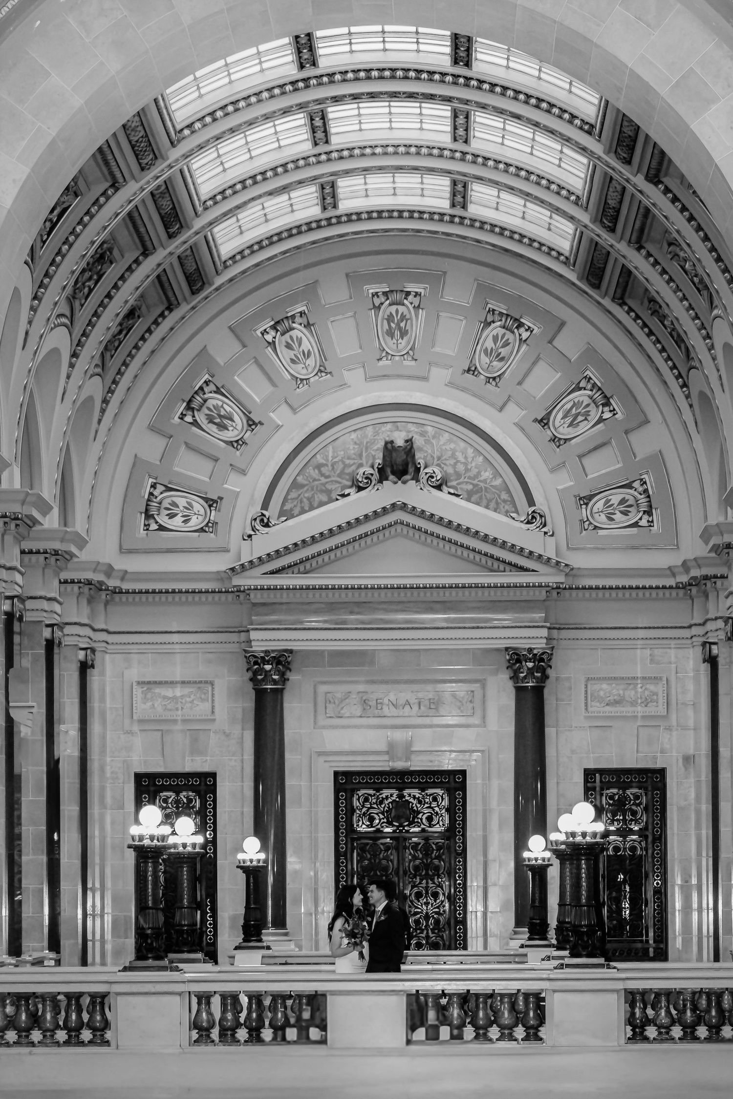Black and white photo of a couple standing before ornate Senate chamber doors in a grand neoclassical building.
