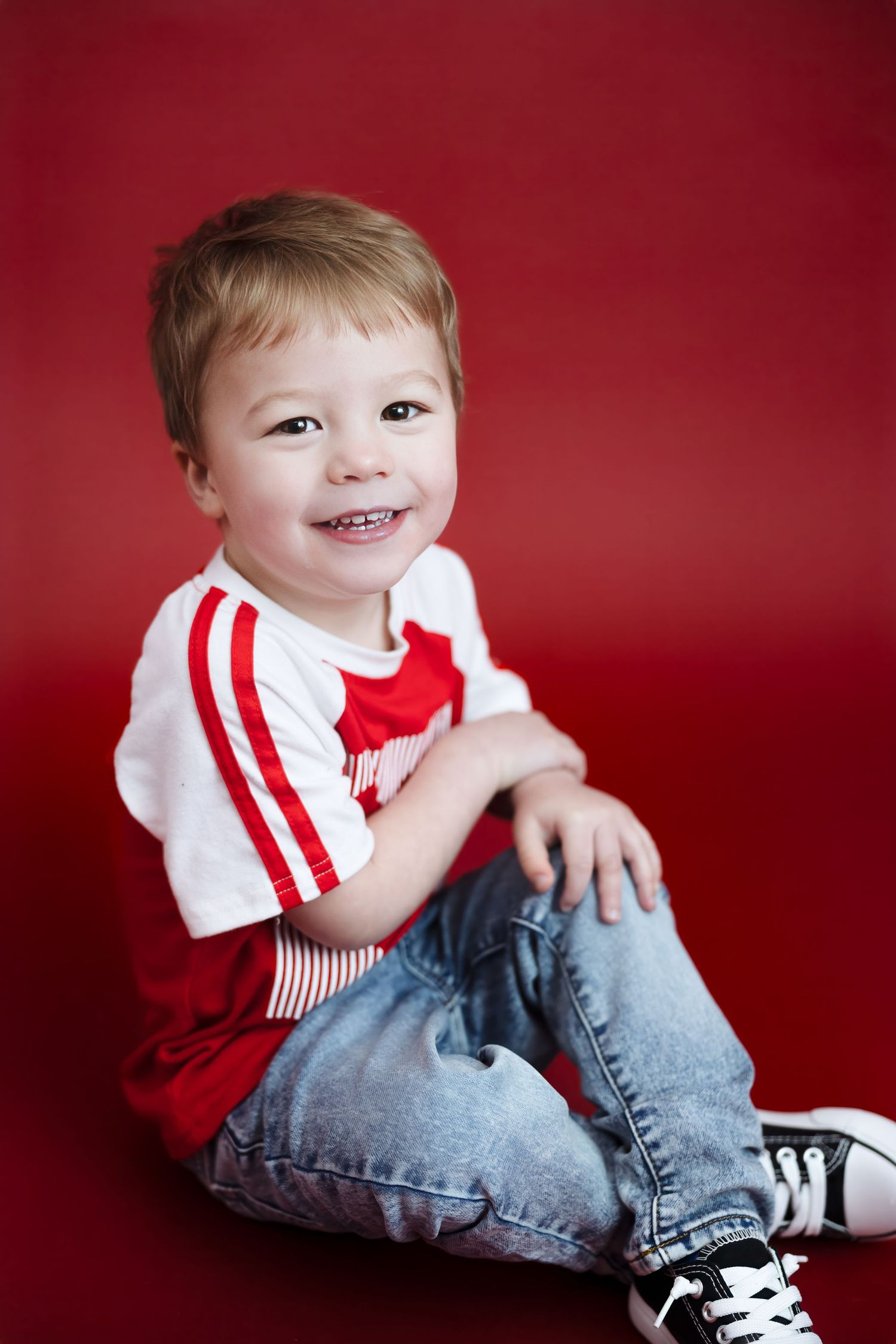 Cheerful toddler in red striped shirt sits smiling on red background.