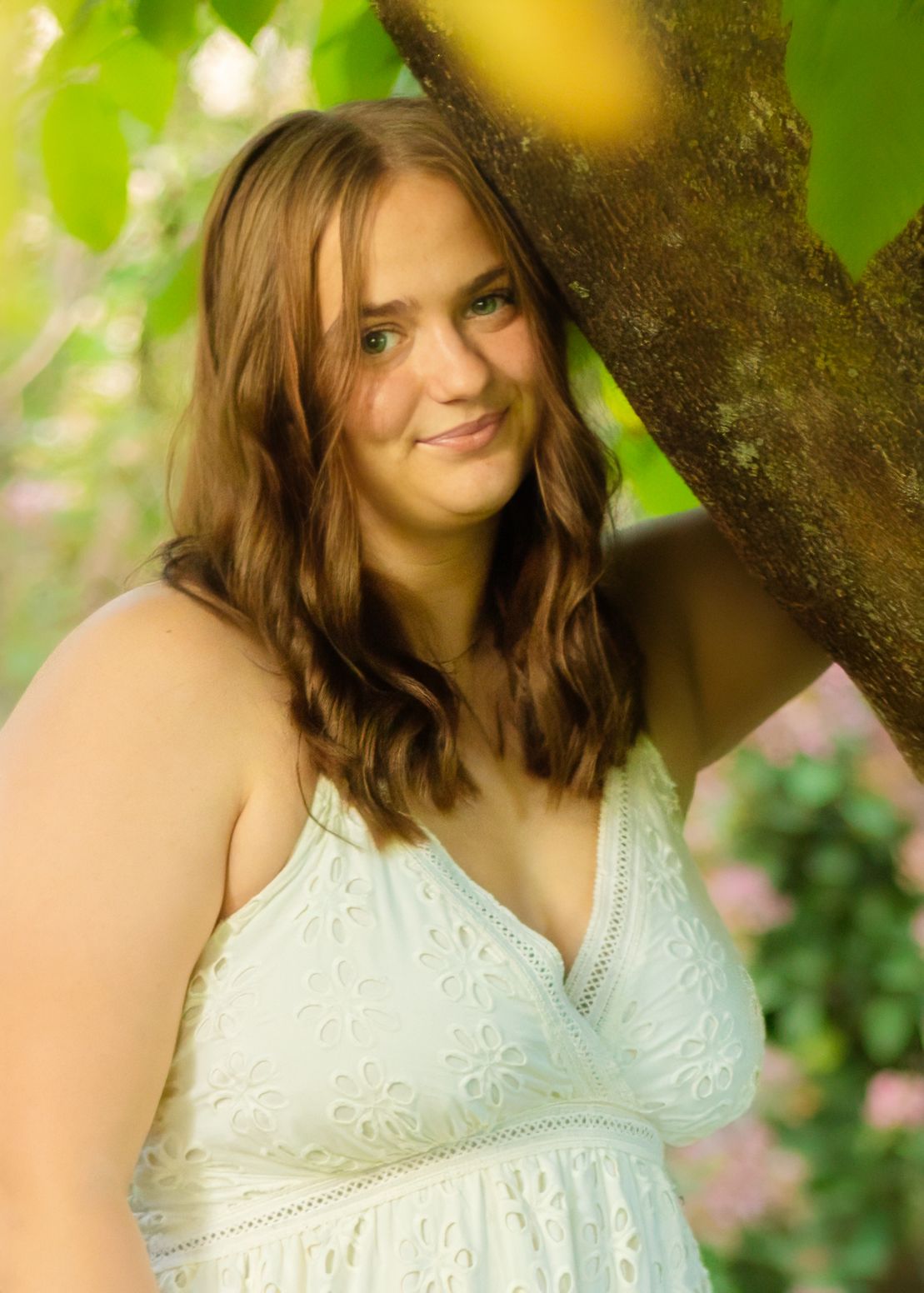 Young woman in white lace dress leaning against tree in blooming garden for senior portrait.