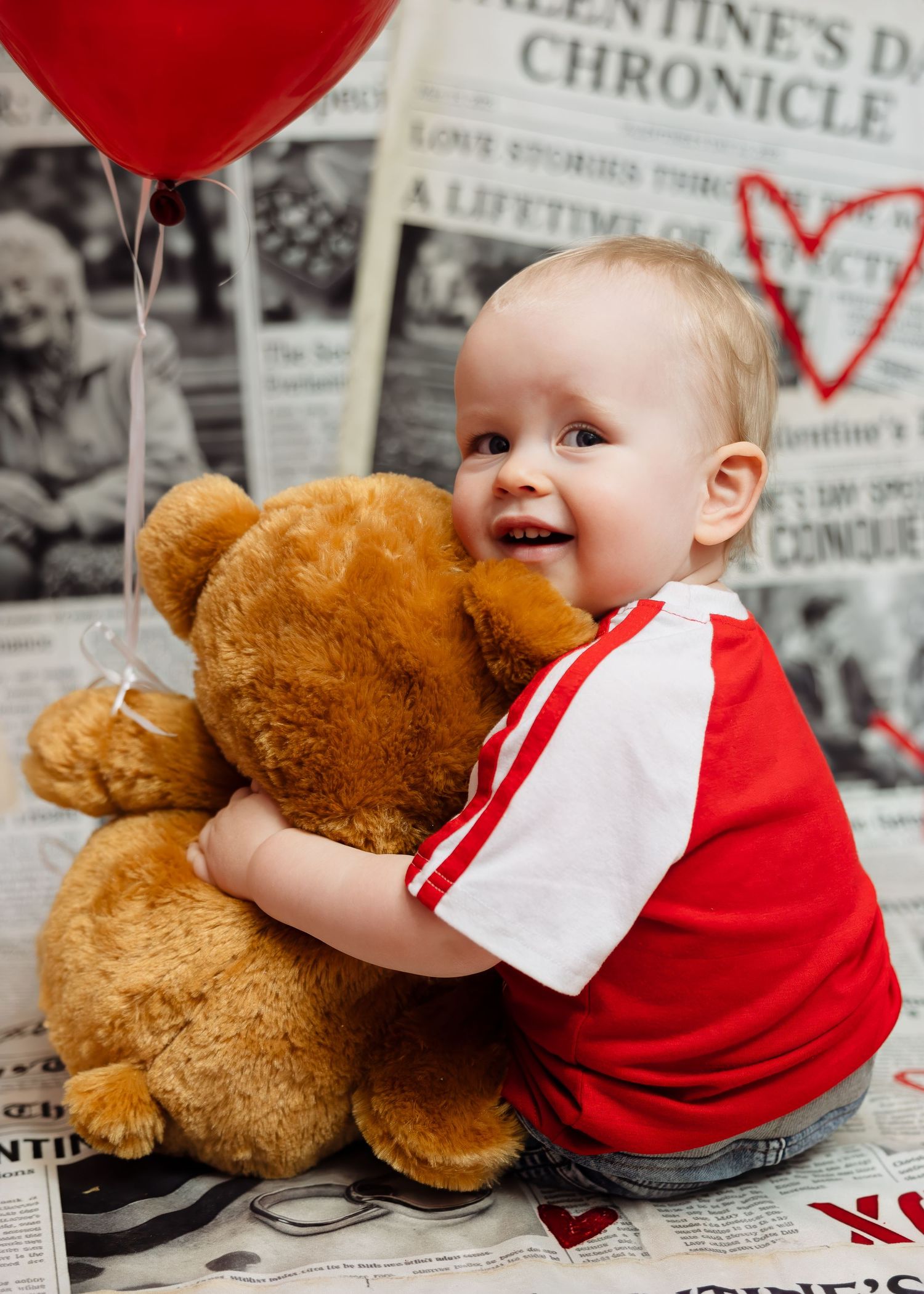 Happy baby hugs large teddy bear with red heart balloon on newspaper backdrop.