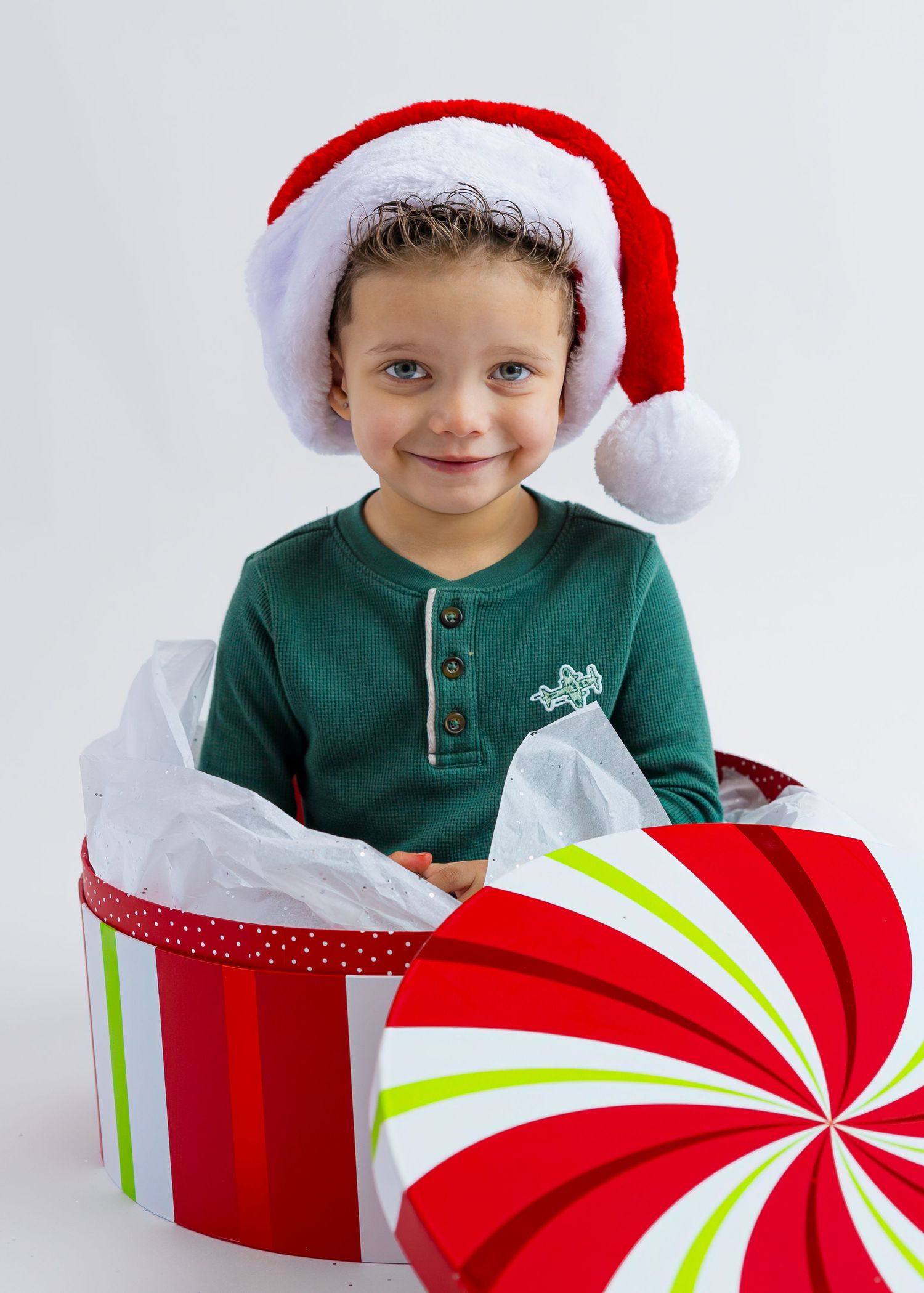 Smiling child wearing Santa hat sitting inside large red and white striped Christmas gift box.