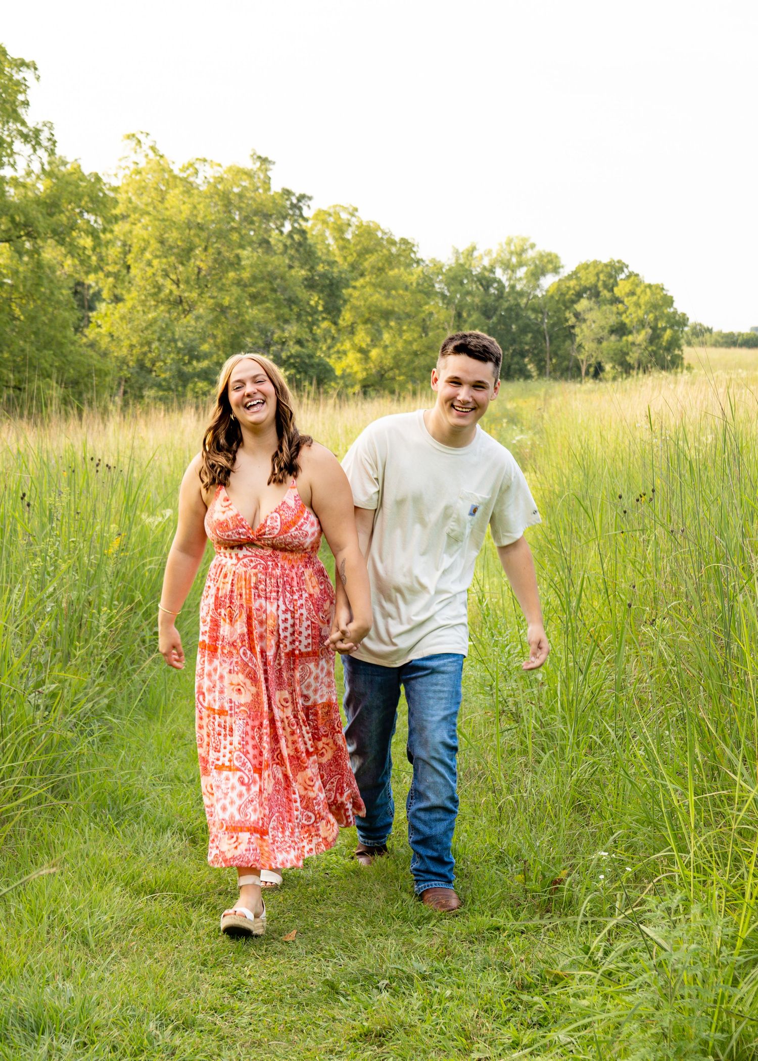 A happy couple walks hand-in-hand through a lush green meadow path, smiling and laughing together.