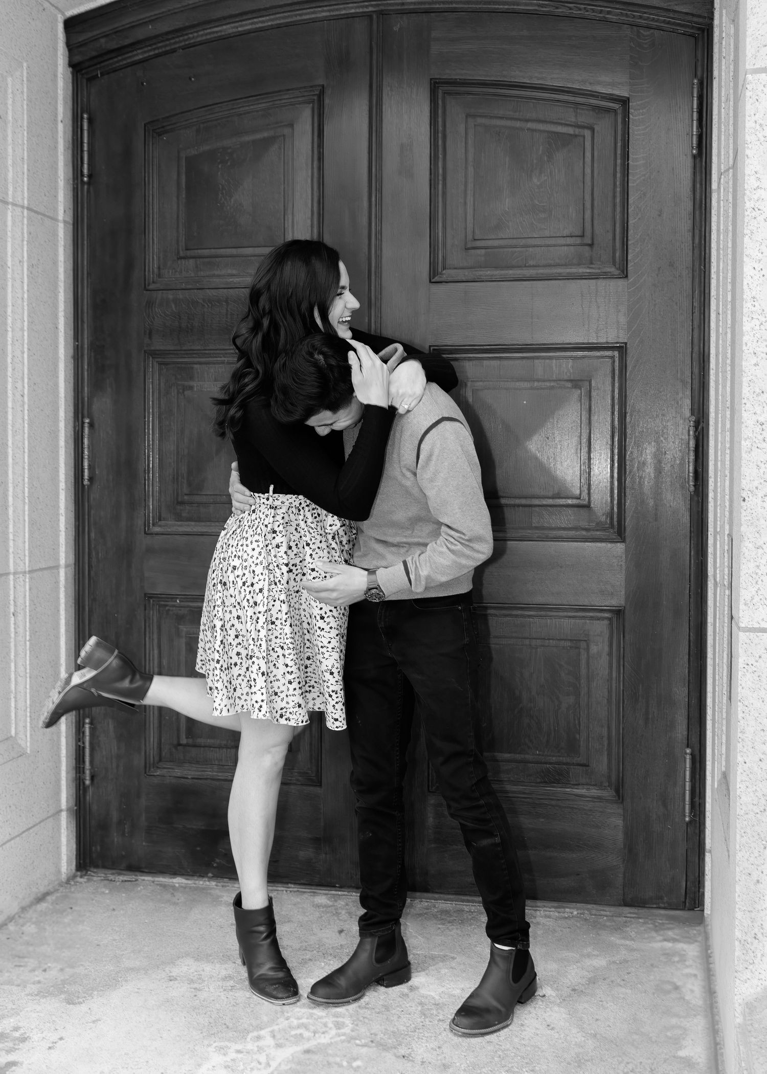 Black and white image of a couple sharing a playful moment in front of large wooden double doors.