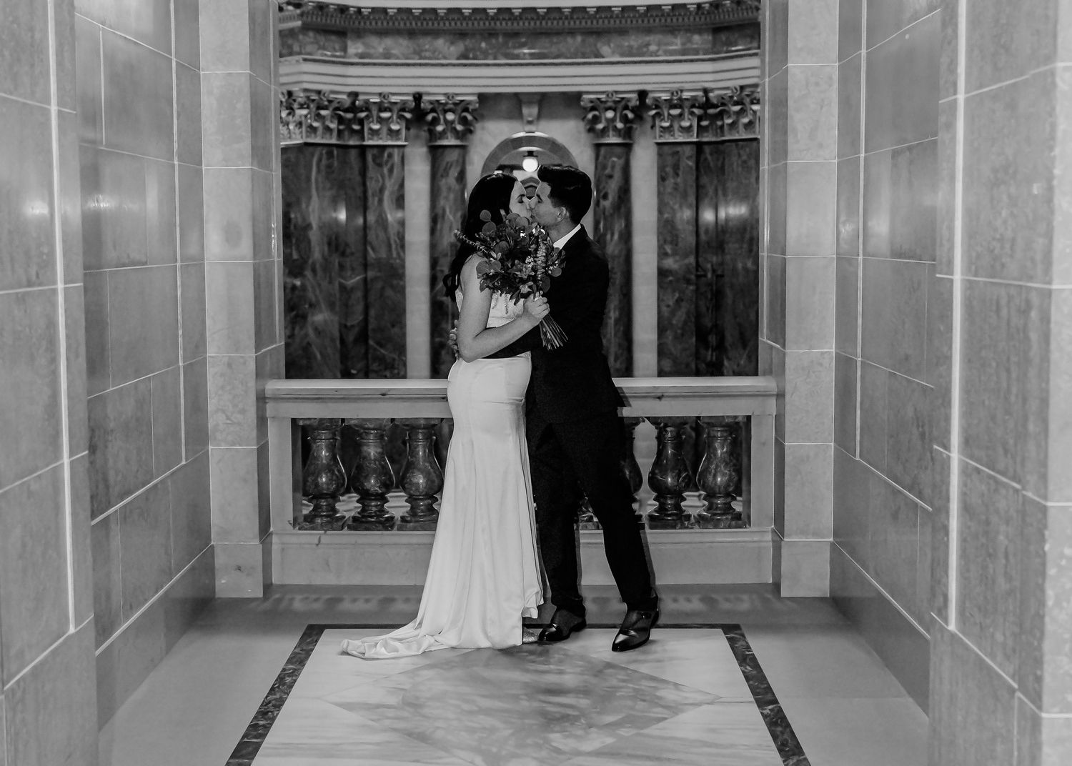 Black and white close-up of bride and groom sharing a kiss in an ornate marble archway on wedding day.