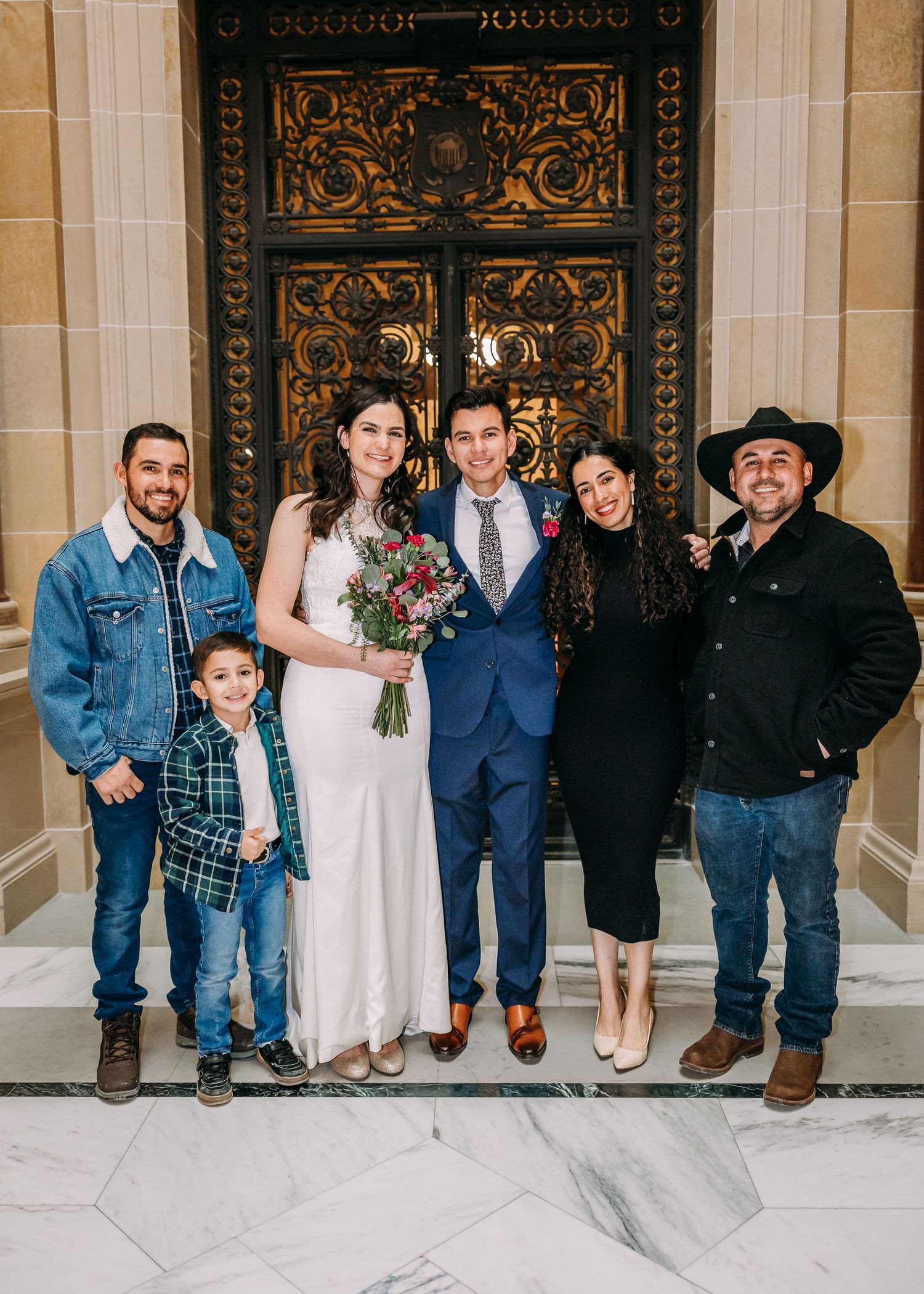 Bride holding bouquet stands with groom and four guests before grand golden doors at a city hall wedding.