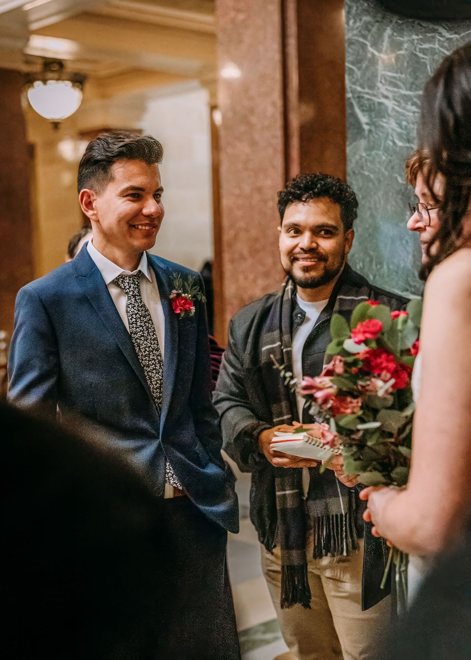 Smiling groom in navy suit chats with guest holding red floral bouquet at elegant wedding venue.