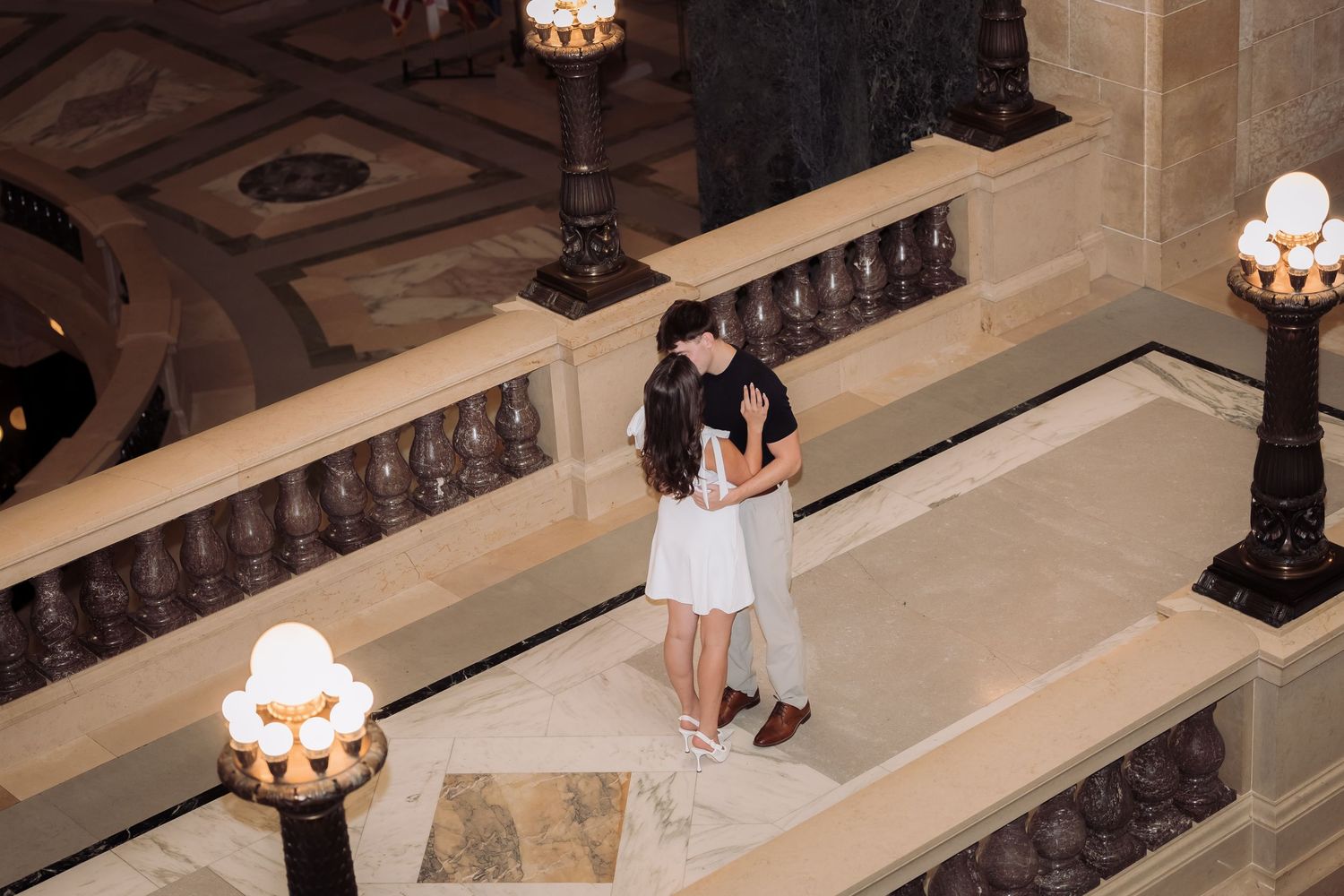 Couple sharing a romantic kiss on a grand marble balcony inside an elegant building with ornate lamp posts.