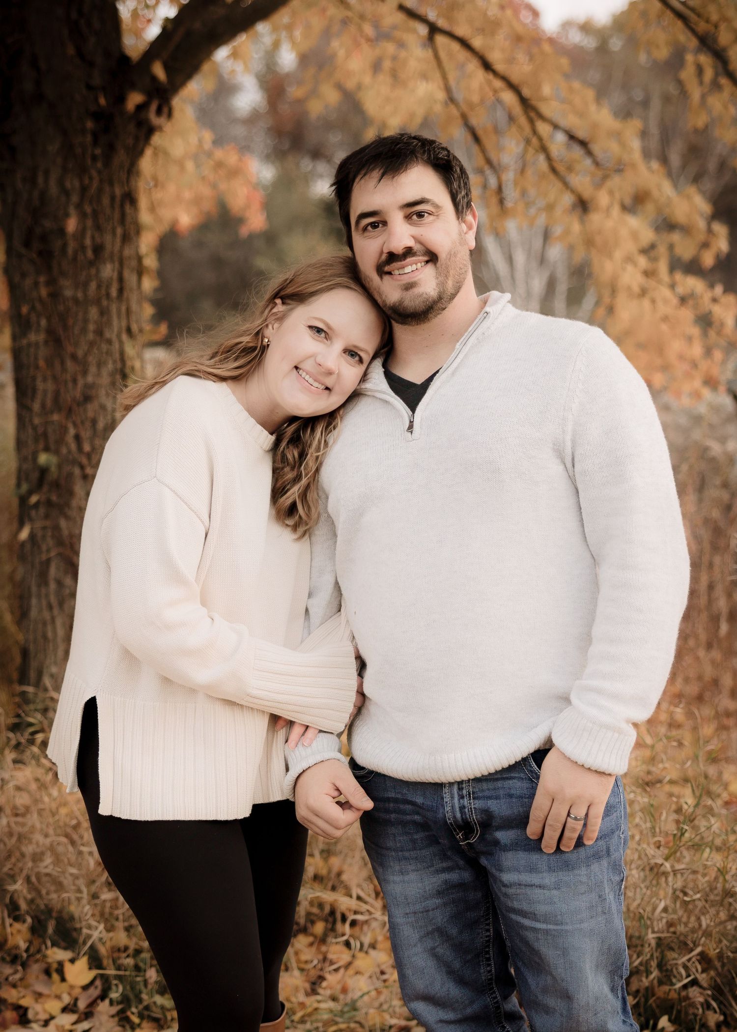A smiling couple poses together under autumn foliage, wearing matching cream sweaters in a fall outdoor setting.