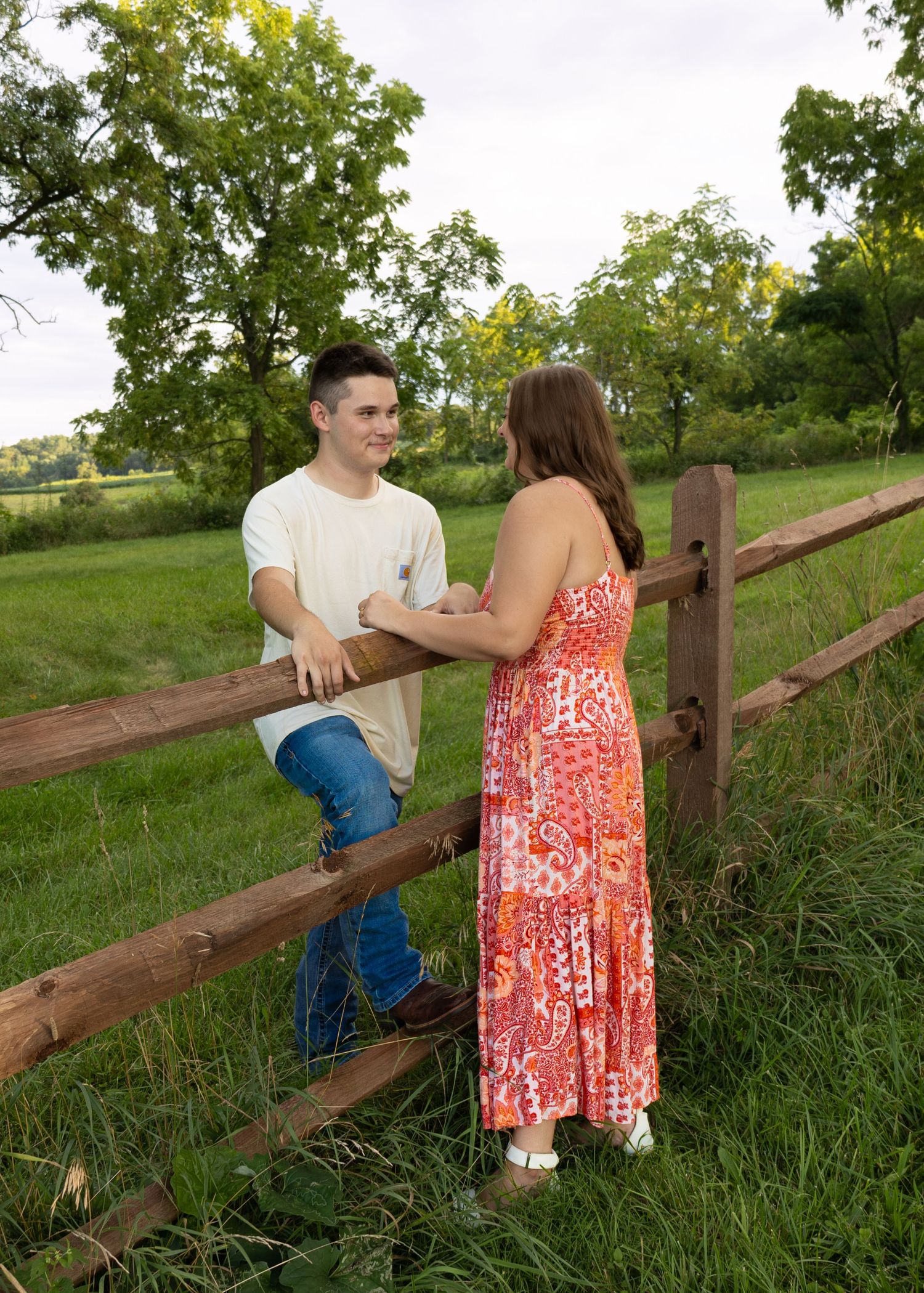 Young couple smiling and chatting by rustic wooden fence in lush green countryside setting.