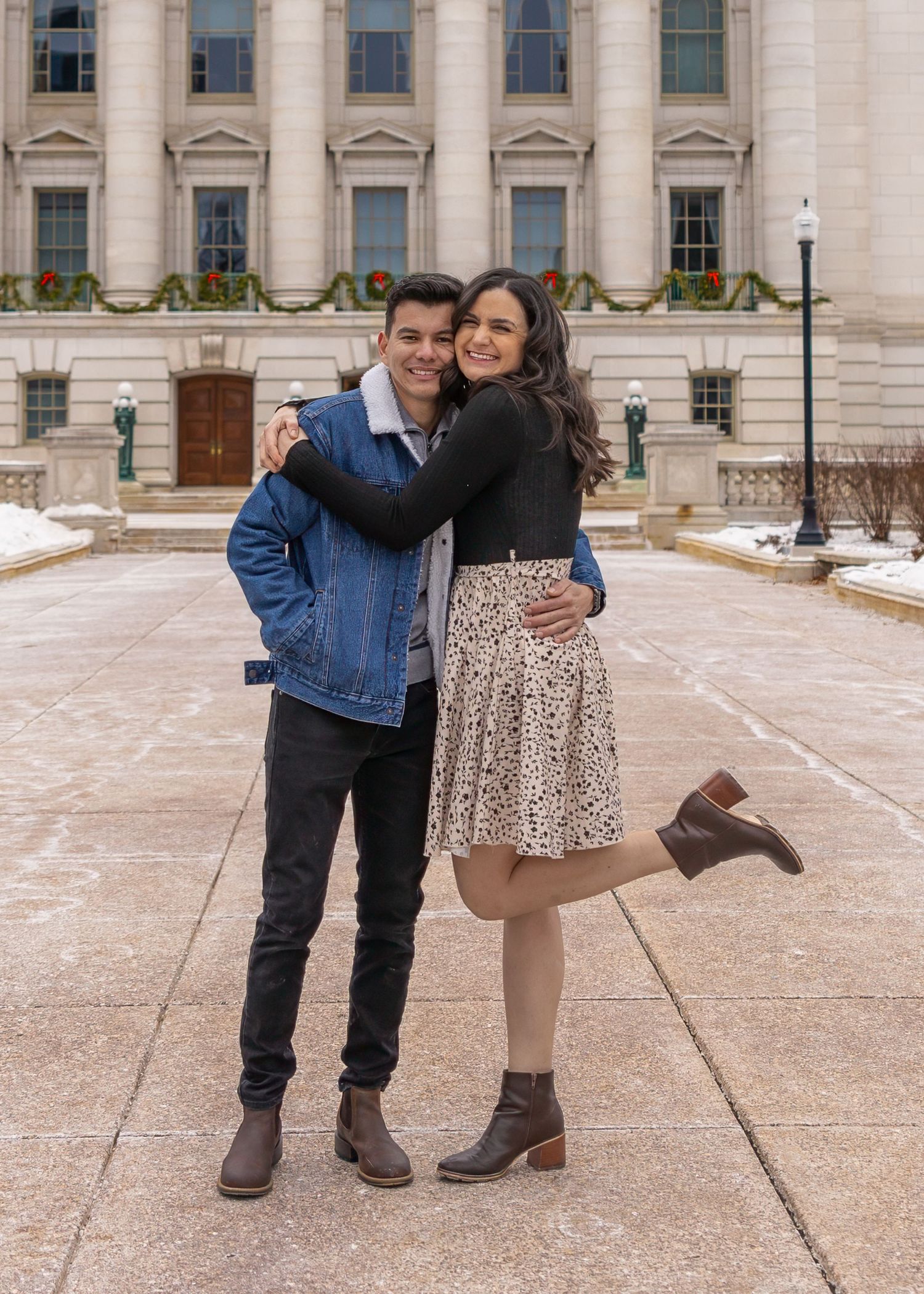 A smiling couple hugs playfully outside a neoclassical building decorated with holiday wreaths in winter.
