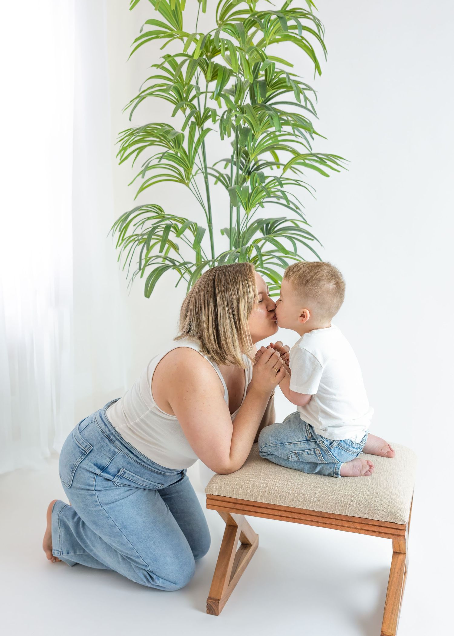Blonde mother kneeling to kiss toddler son seated on bench beside tall green indoor plant.