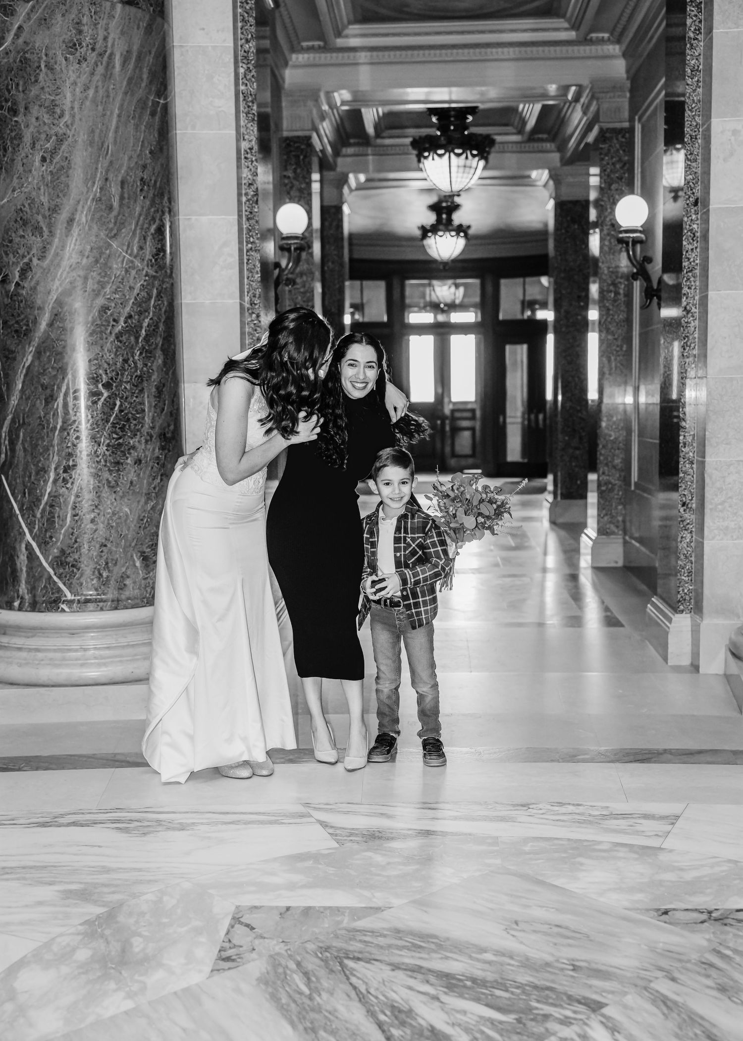 Bride in white gown poses with woman in black dress and young boy holding flowers in elegant marble hall.