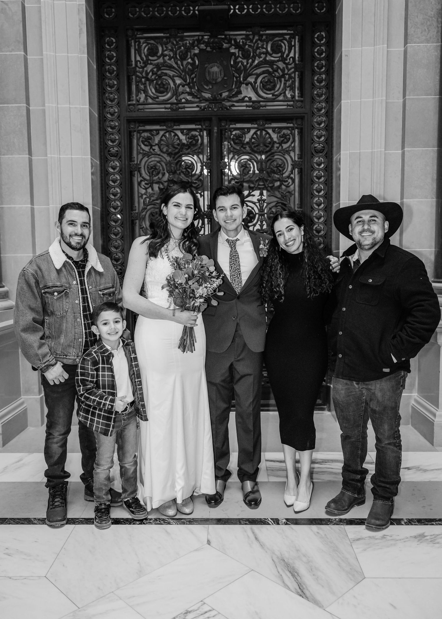 Black and white photo of bride, groom, and five guests posing before an ornate door at a city hall wedding.