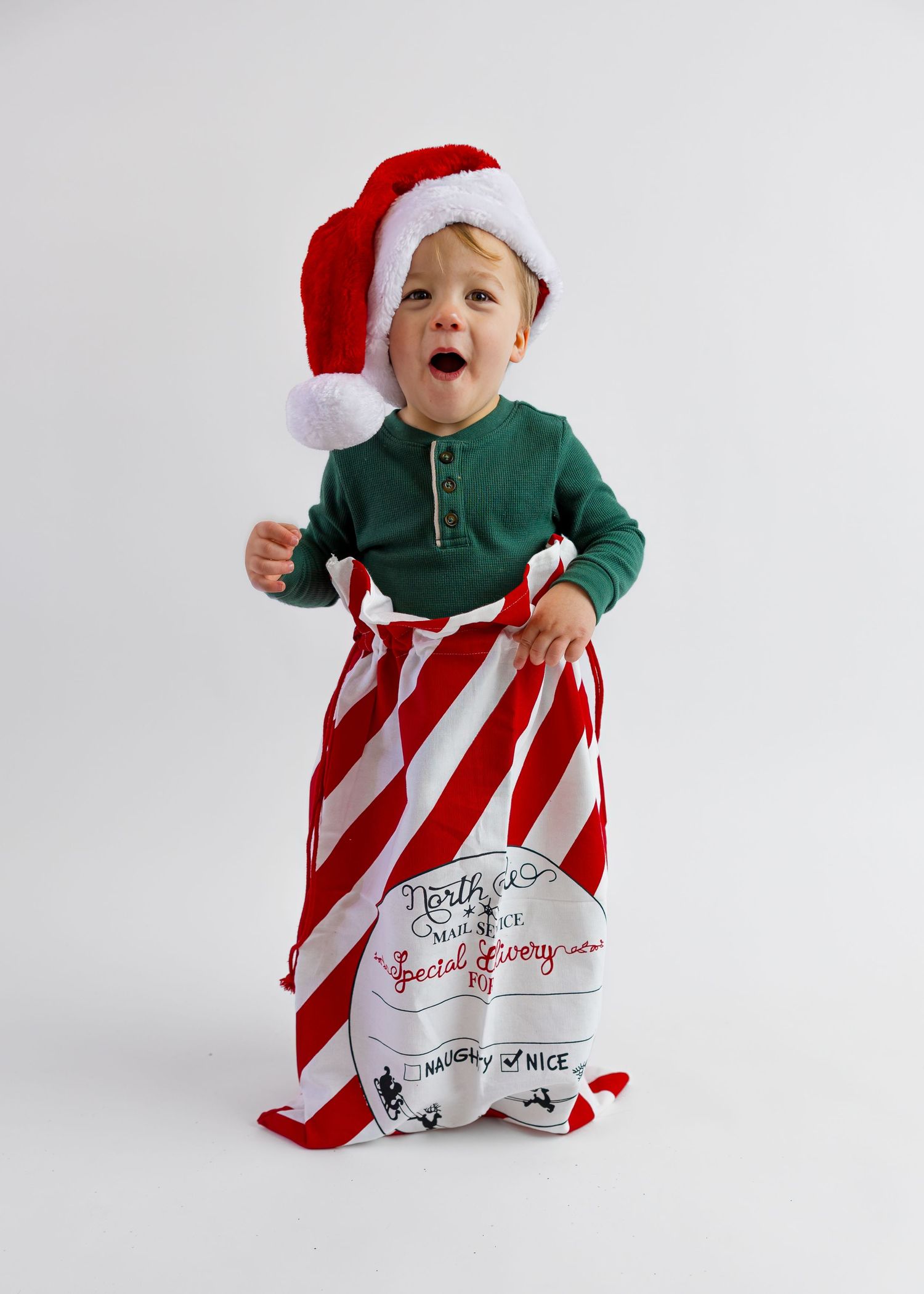 Smiling baby in green henley and Santa hat holding a red and white striped Christmas sack.