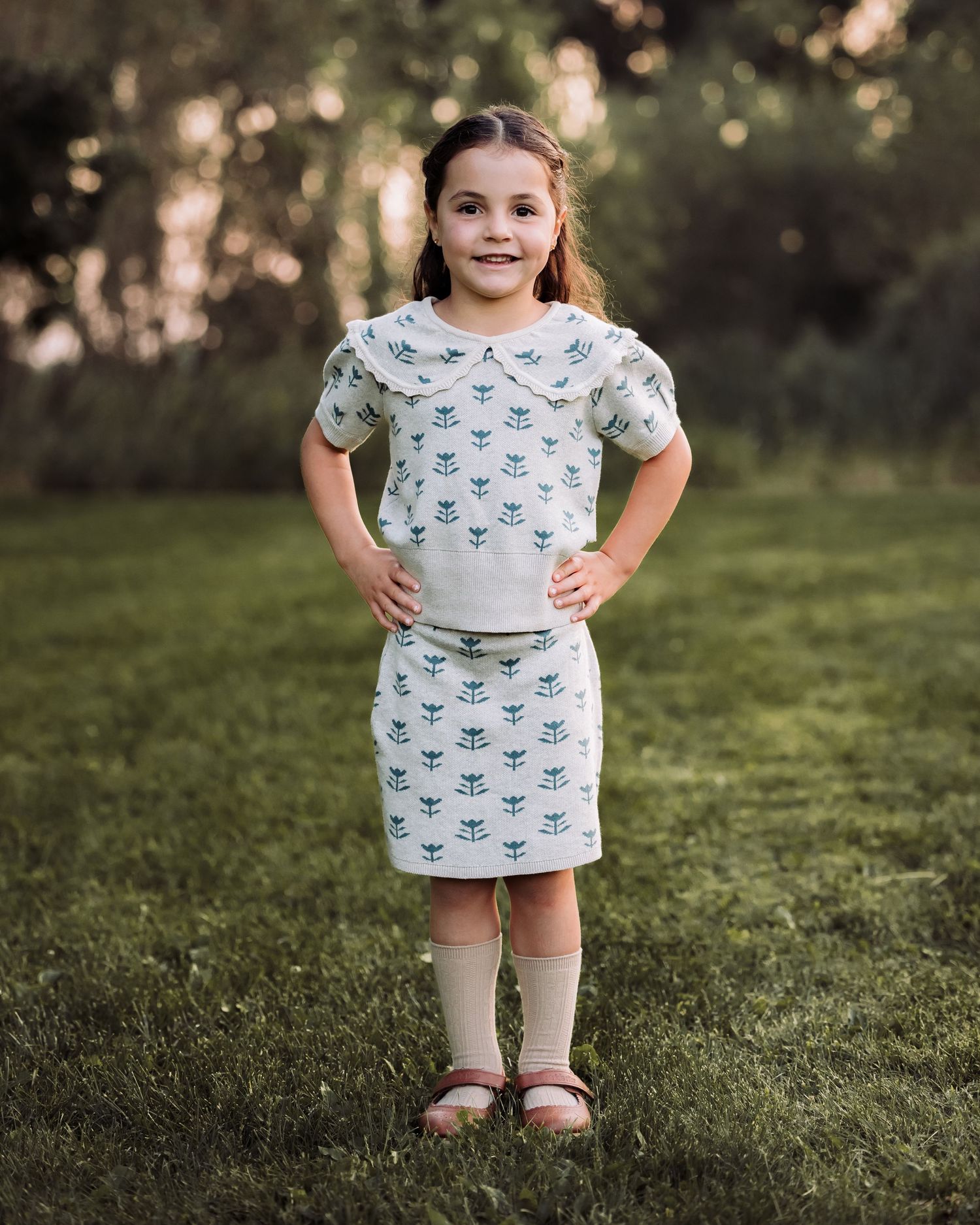 Young girl in matching floral print top and skirt set standing on green grass outdoors.
