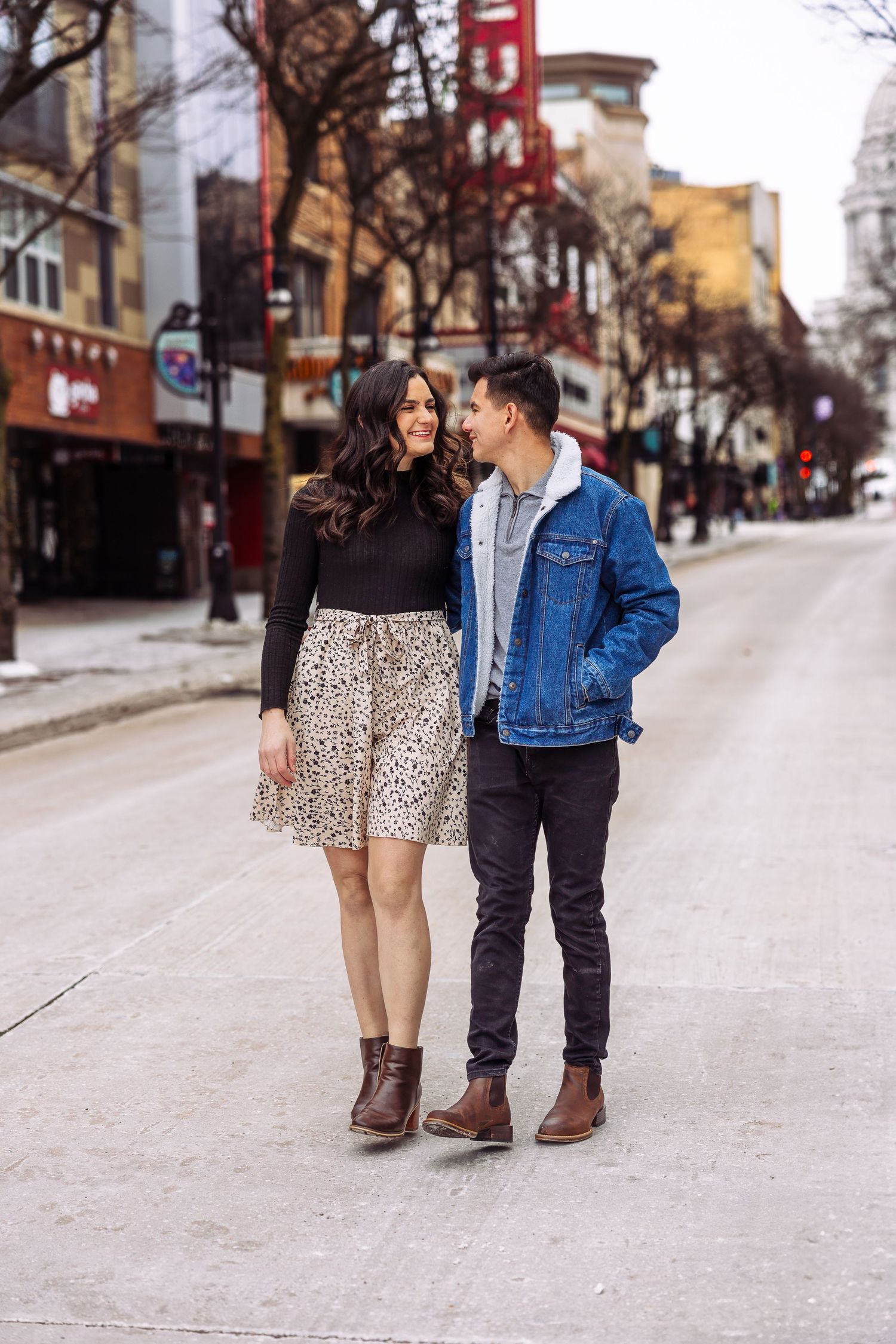 Couple smiling at each other while walking on a city street, woman in floral skirt and man in denim jacket.