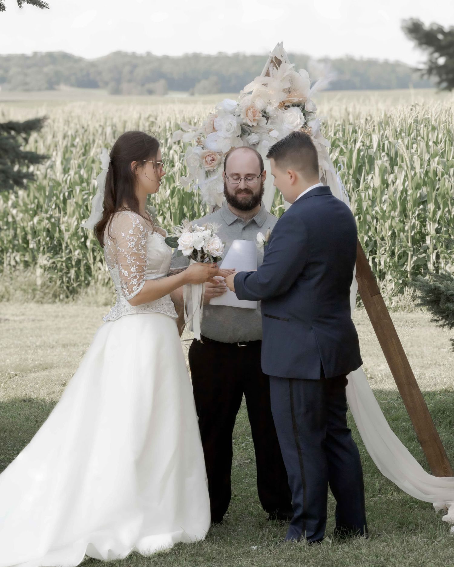 Bride and groom exchange vows outdoors before a floral arch with a cornfield backdrop.