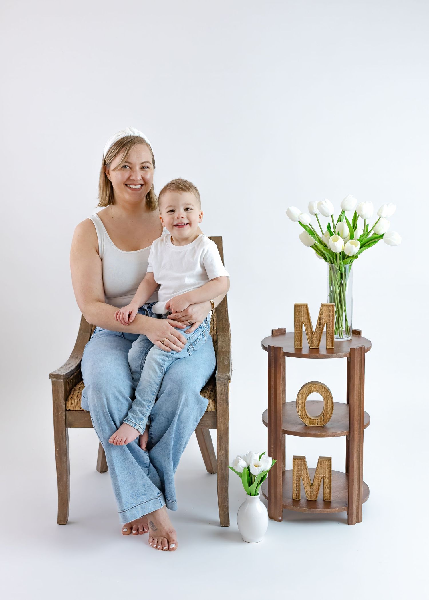 Smiling blonde mother sitting in wooden chair holding happy toddler son beside MOM letter decor.
