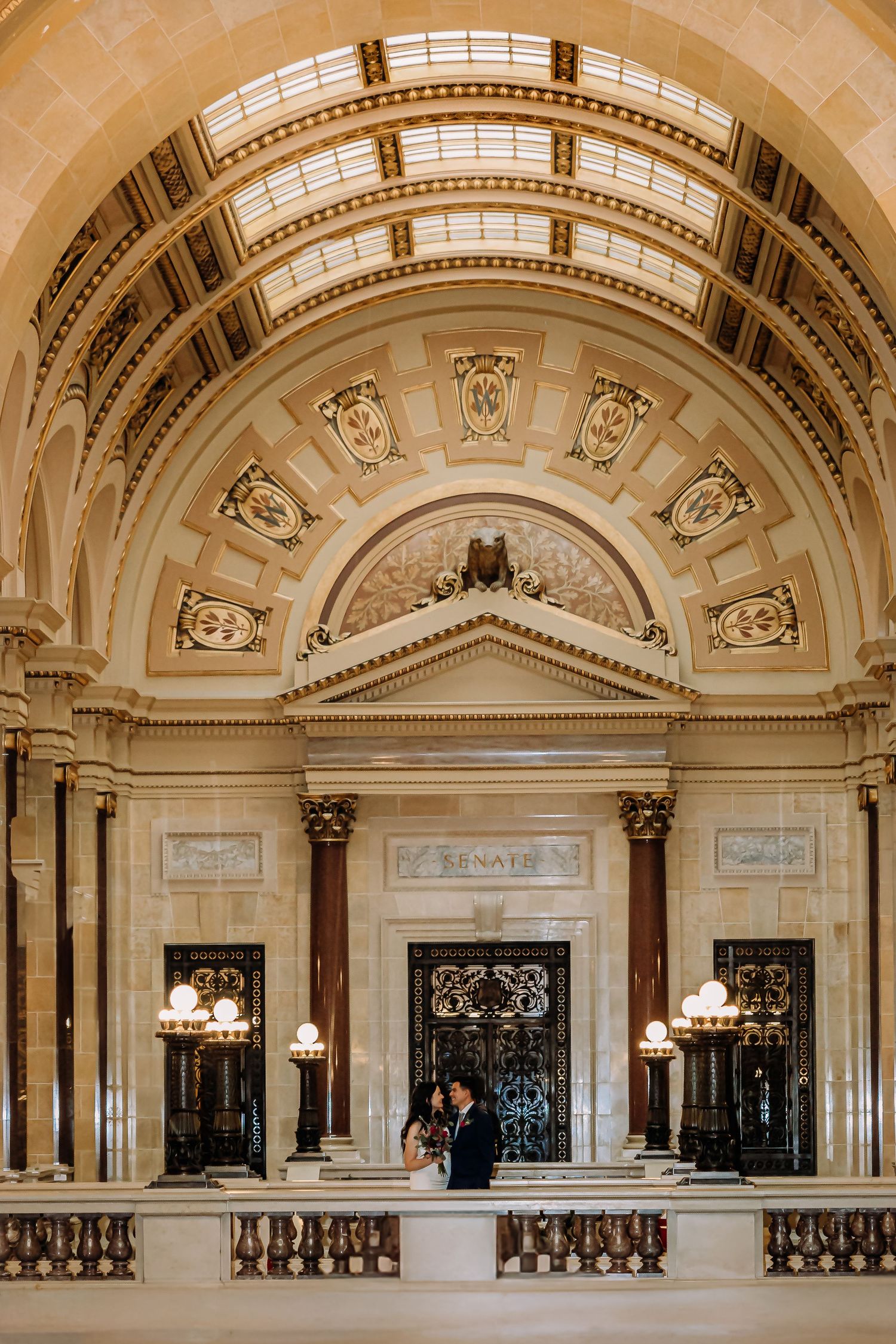 Bride and groom stand beneath ornate arched ceiling in grand neoclassical building interior.