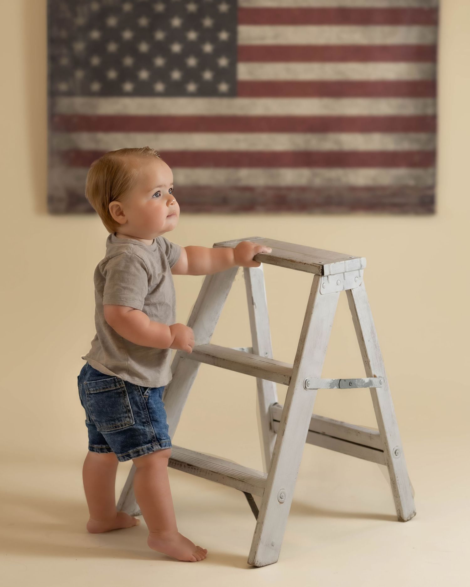 Baby boy in gray shirt and denim shorts stands holding a small wooden ladder before an American flag.