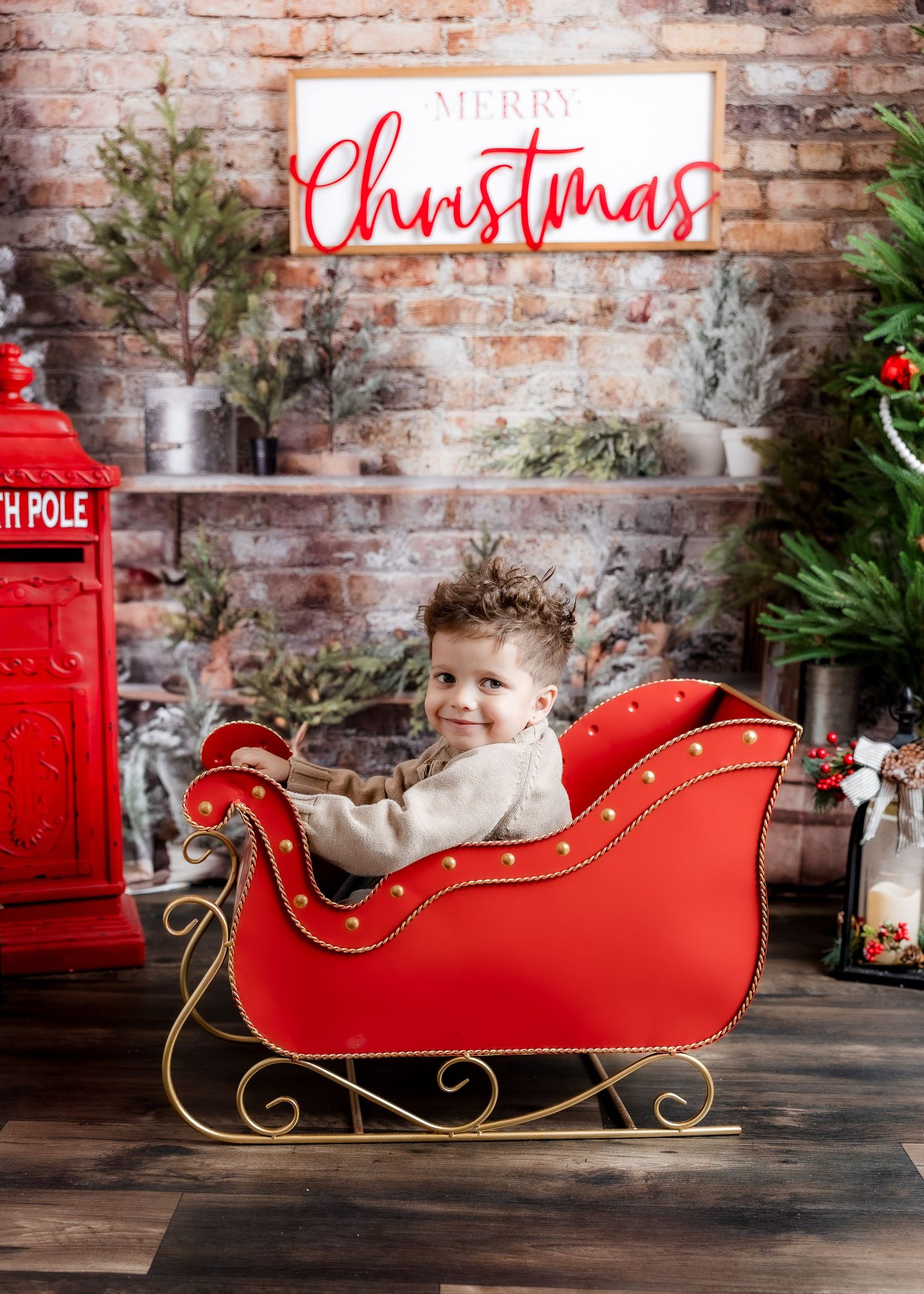 Toddler boy sitting in a red Christmas sleigh with festive holiday decorations and Merry Christmas sign.