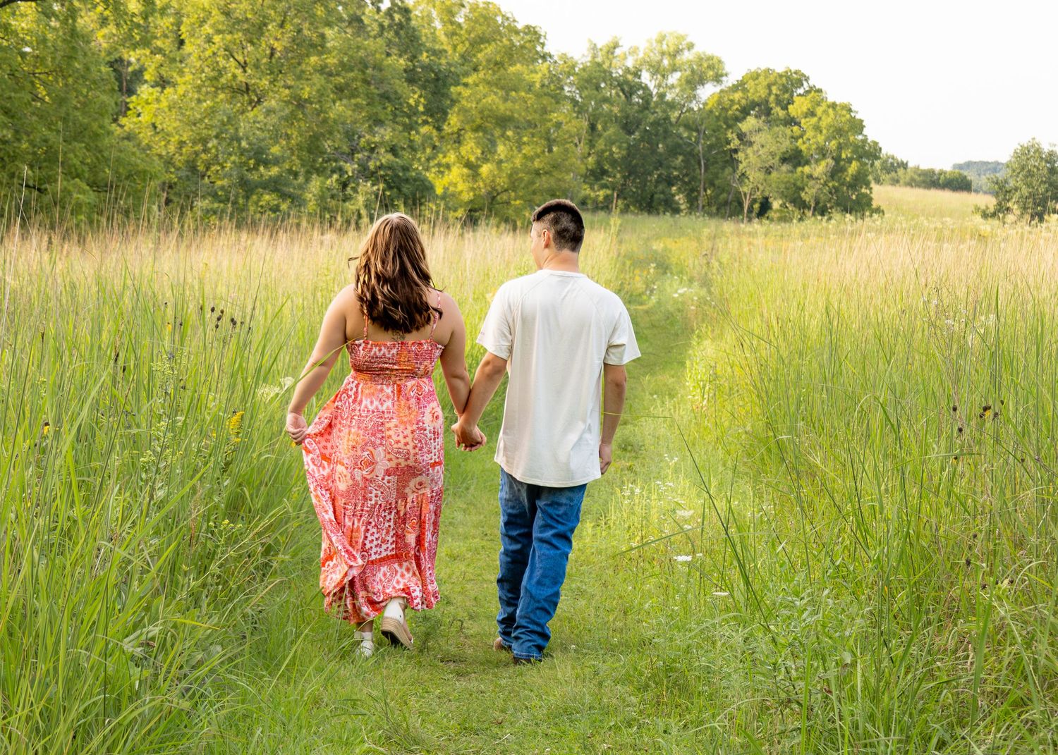 A couple walks hand-in-hand away from the camera along a lush green meadow trail in summer.