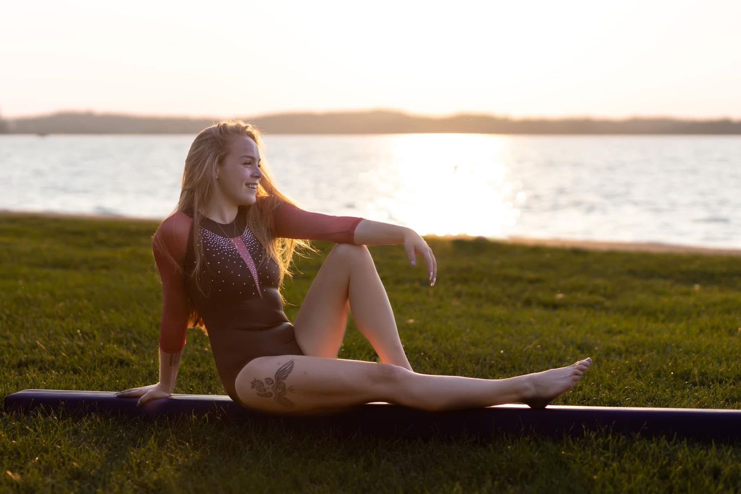 A young blonde woman sits on a mat by a lake at sunset, gazing peacefully at the water.