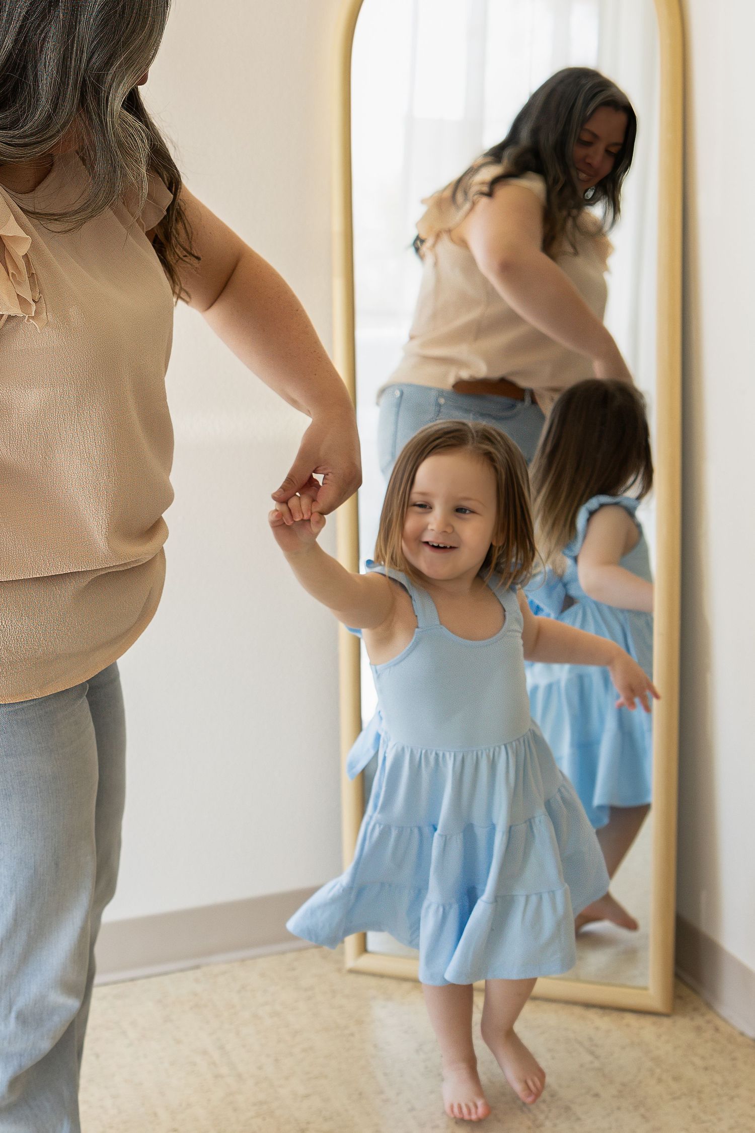 A happy toddler girl in a blue tiered dress dances barefoot while holding her mothers hand near a mirror.