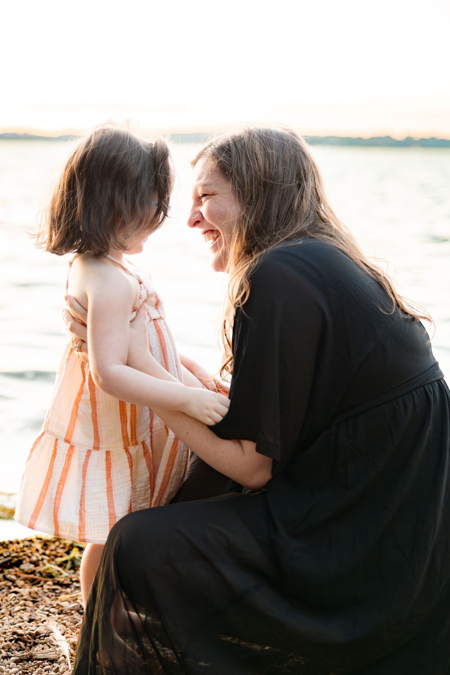 A mother in a black dress crouches by the water, smiling at her young daughter in a pink striped dress at sunset.