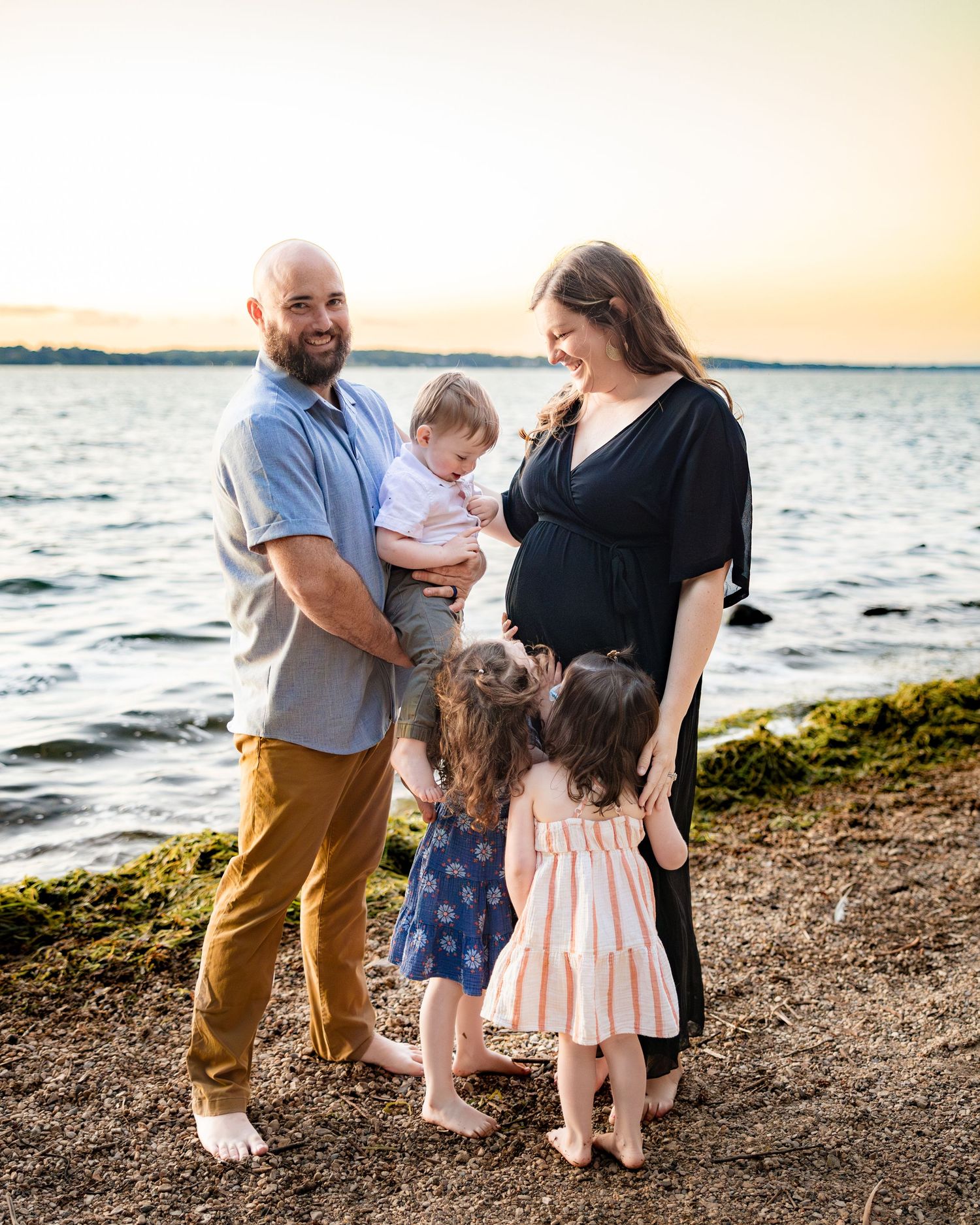 Pregnant woman and bald man with two young girls stand barefoot on a rocky beach at golden sunset.
