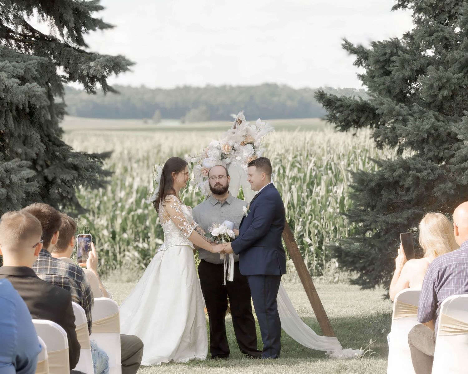 Outdoor farm wedding ceremony with bride in white gown and groom in navy suit before a cornfield backdrop.